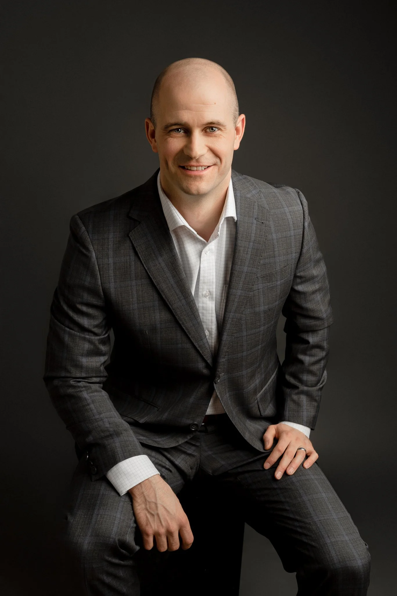 A man in a checkered suit sitting against a dark background, smiling at the camera.