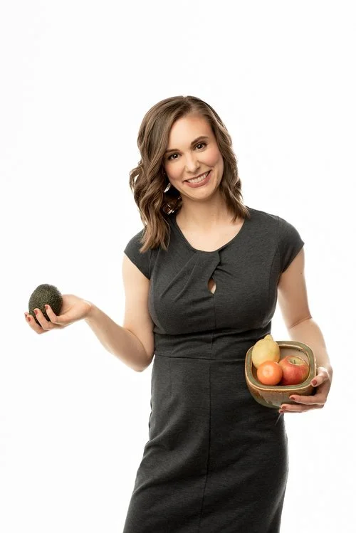 Smiling woman in a black dress holding a bowl of apples and a small dark fruit in her hands against a white background.