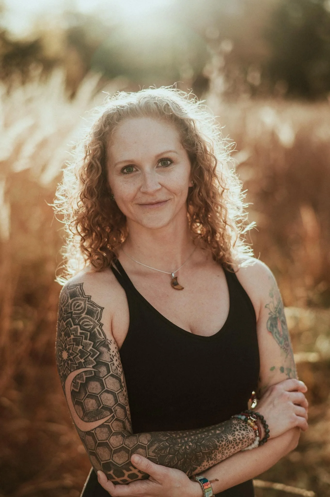 A woman with curly red hair and tattoos on her arms, wearing a black sleeveless top and jewelry, standing outdoors during sunset in a field of tall grass.