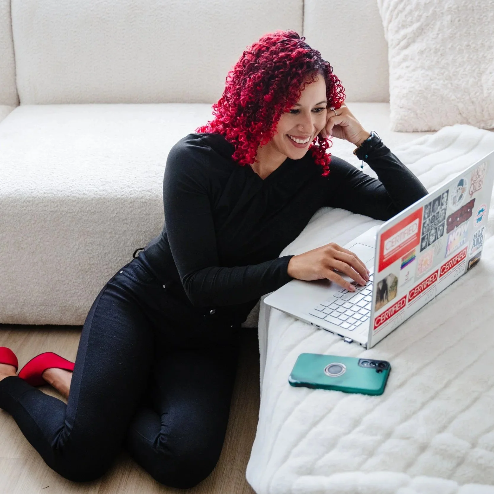 A woman with curly red hair is lying on her stomach on the floor, smiling at her laptop which is decorated with stickers. She is wearing a black long-sleeve top, black pants, and red heels, with a large beige couch in the background.