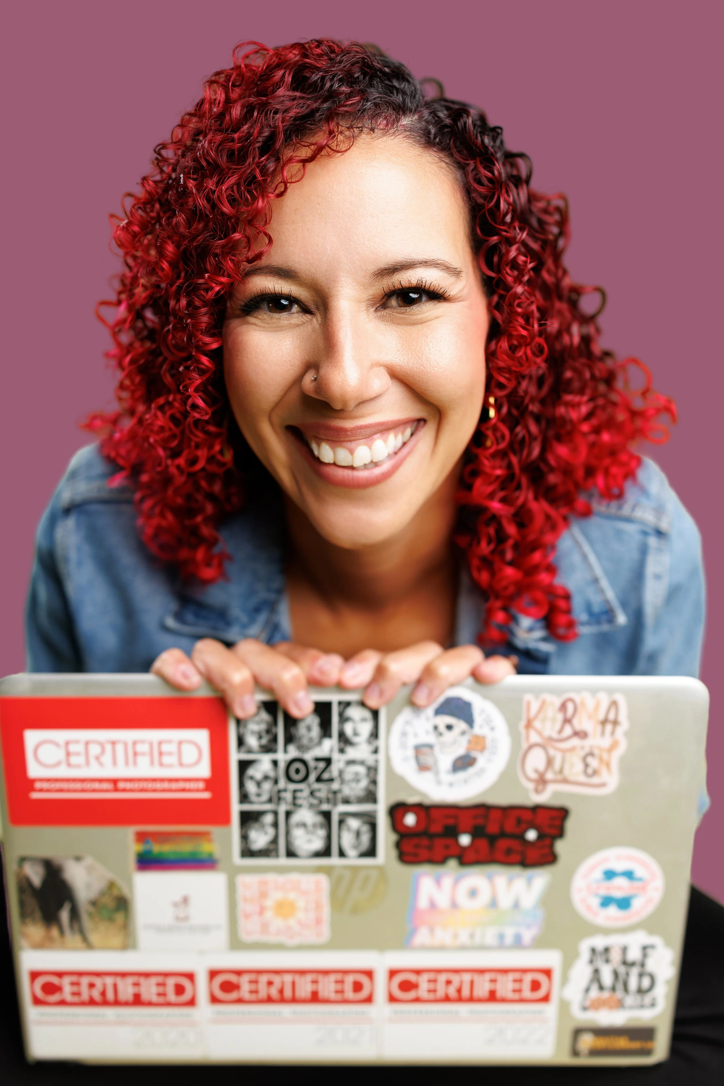 A woman with vibrant red curly hair, smiling, holding a laptop covered with various stickers, against a pink background.