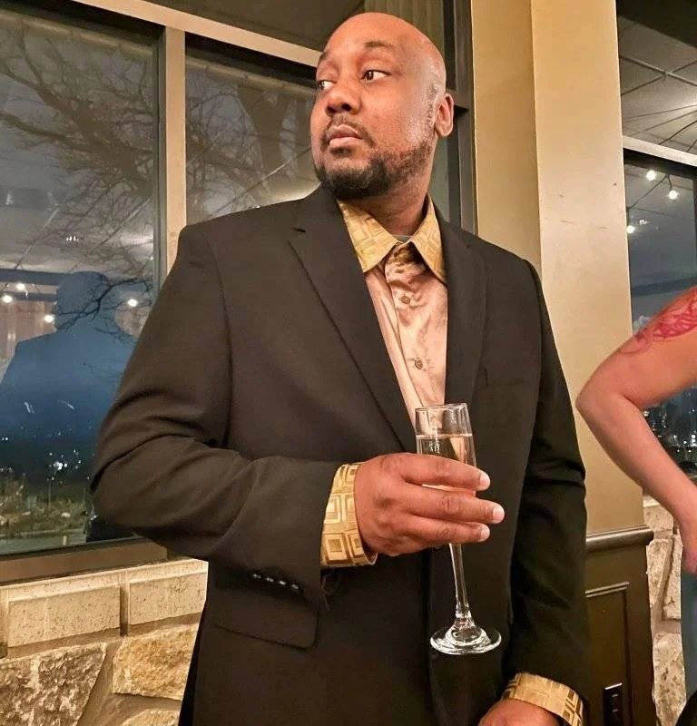 A man dressed in a black suit holding a glass of champagne at a social gathering in an indoor venue with large windows and city view at night.
