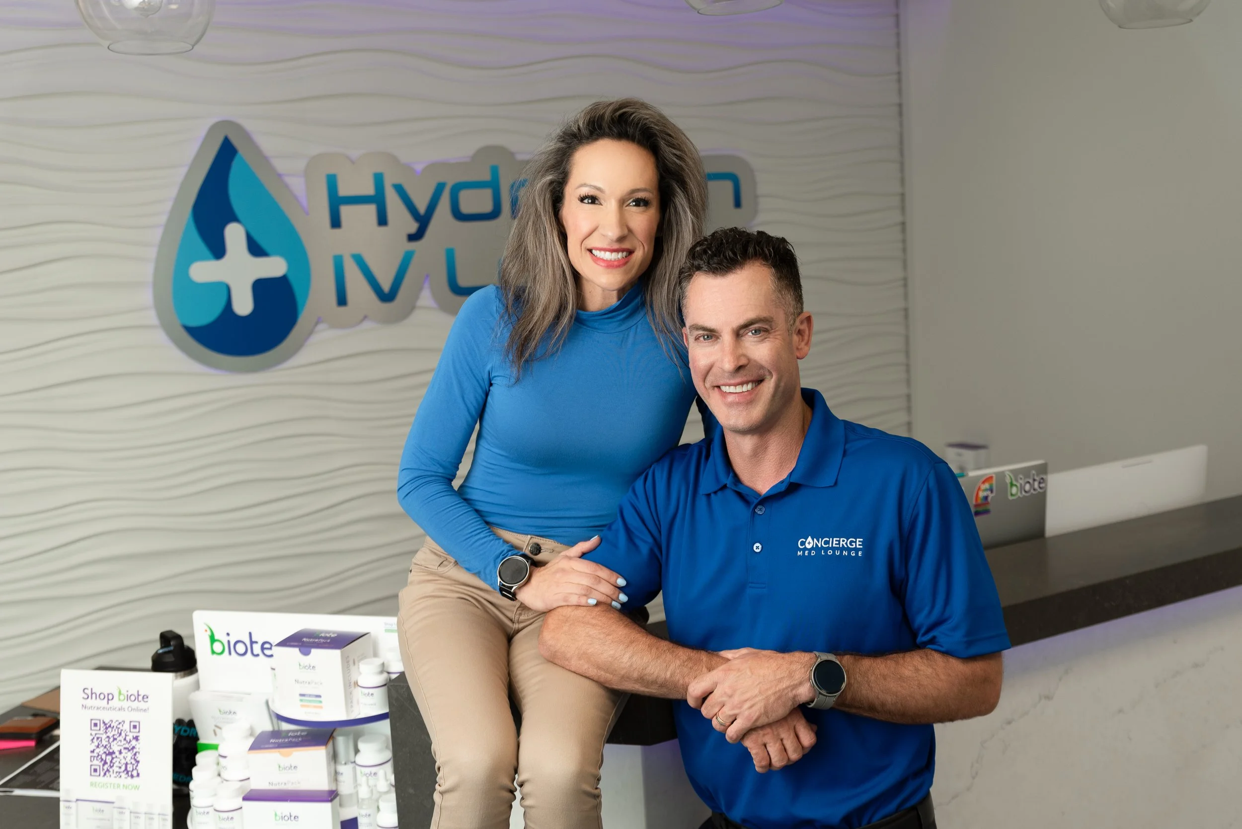 A woman and a man smiling at the camera at a reception desk. The woman is sitting on the desk, wearing a blue long-sleeved top and beige pants, with a smartwatch. The man is standing next to her, wearing a blue polo shirt with a logo that reads 'Concierge Med Lounge'. On the desk, there are various health and wellness products by Biote, and a sign with a QR code. A large sign in the background has a water droplet with a cross and the words 'Hydration IV Lounge'.