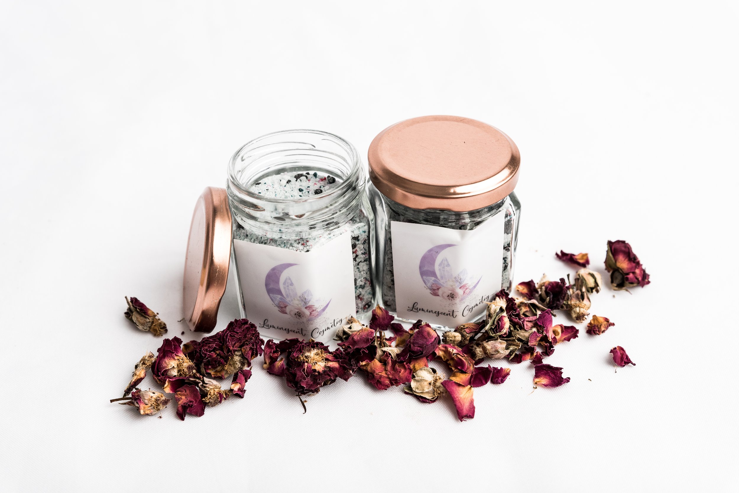 Two glass jars labeled MyLuminous with rose gold lids, one open with sparkly glitter, surrounded by dried rose petals on a white background.