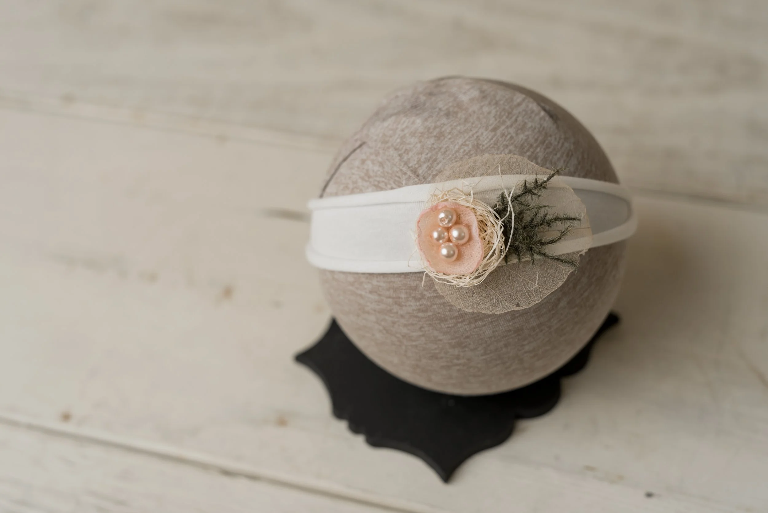 Decorative round gift box wrapped with a white band, adorned with a pink fabric flower, small pearls, and dried greenery, placed on a black scalloped coaster on a white wooden surface.