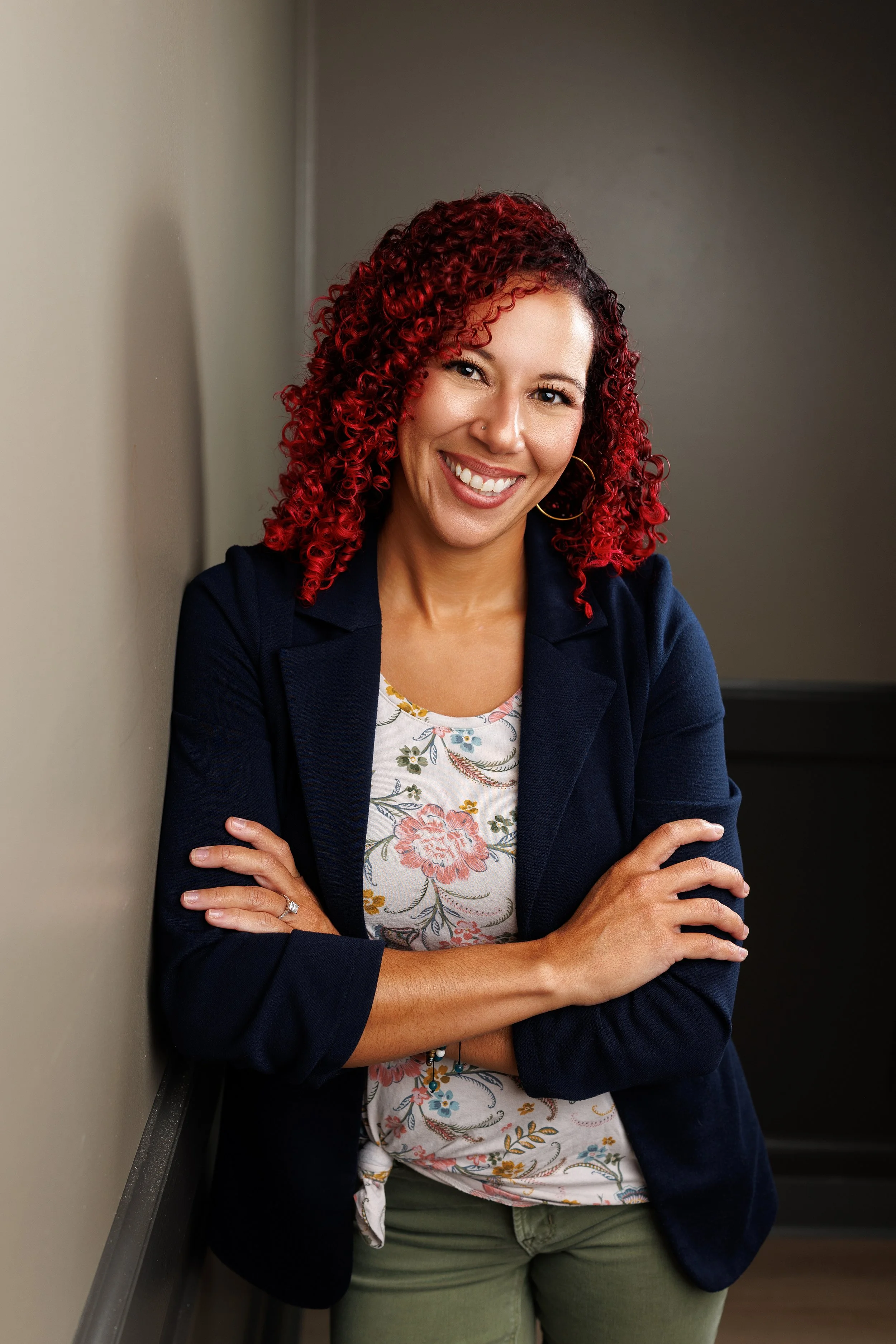 A woman with curly red hair, wearing a navy blazer and a floral shirt, smiling and crossing her arms while leaning against a wall.