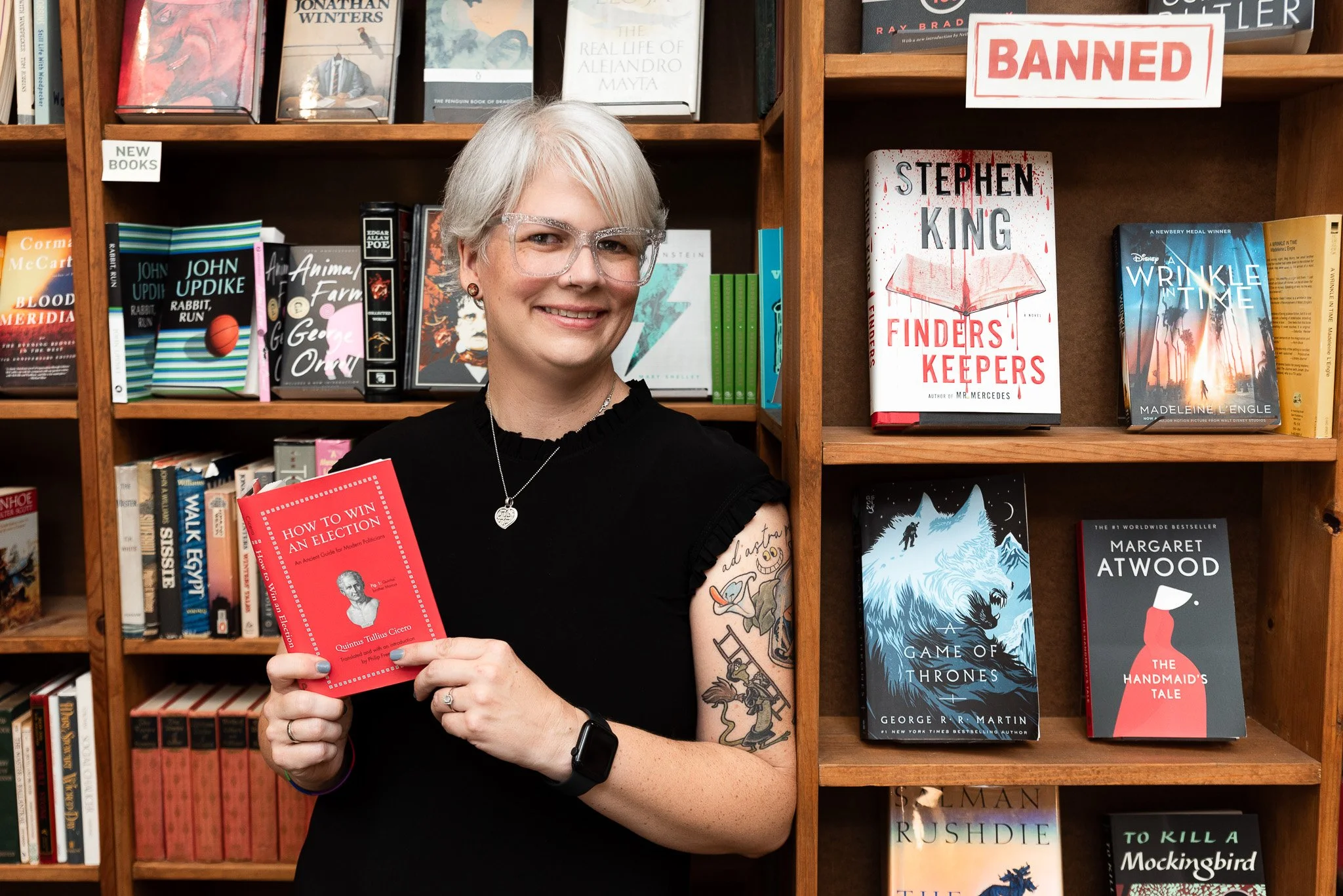 A woman with white hair, glasses, and tattoos on her arm, standing in front of bookshelves in a bookstore, holding a red book titled 'How to Win an Election'.