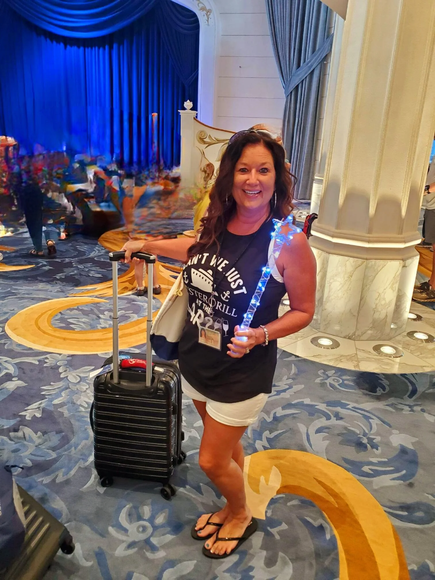 Woman smiling while holding a blue light-up star wand, standing on a cruise ship with luggage, wearing a black shirt and white shorts.