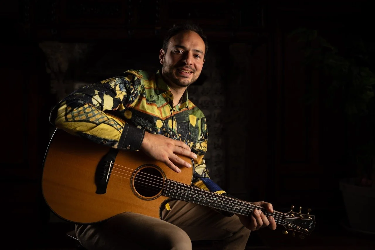 Un homme souriant joue de la guitare en fauteuil dans un environnement sombre avec un mur en pierre en arrière-plan.