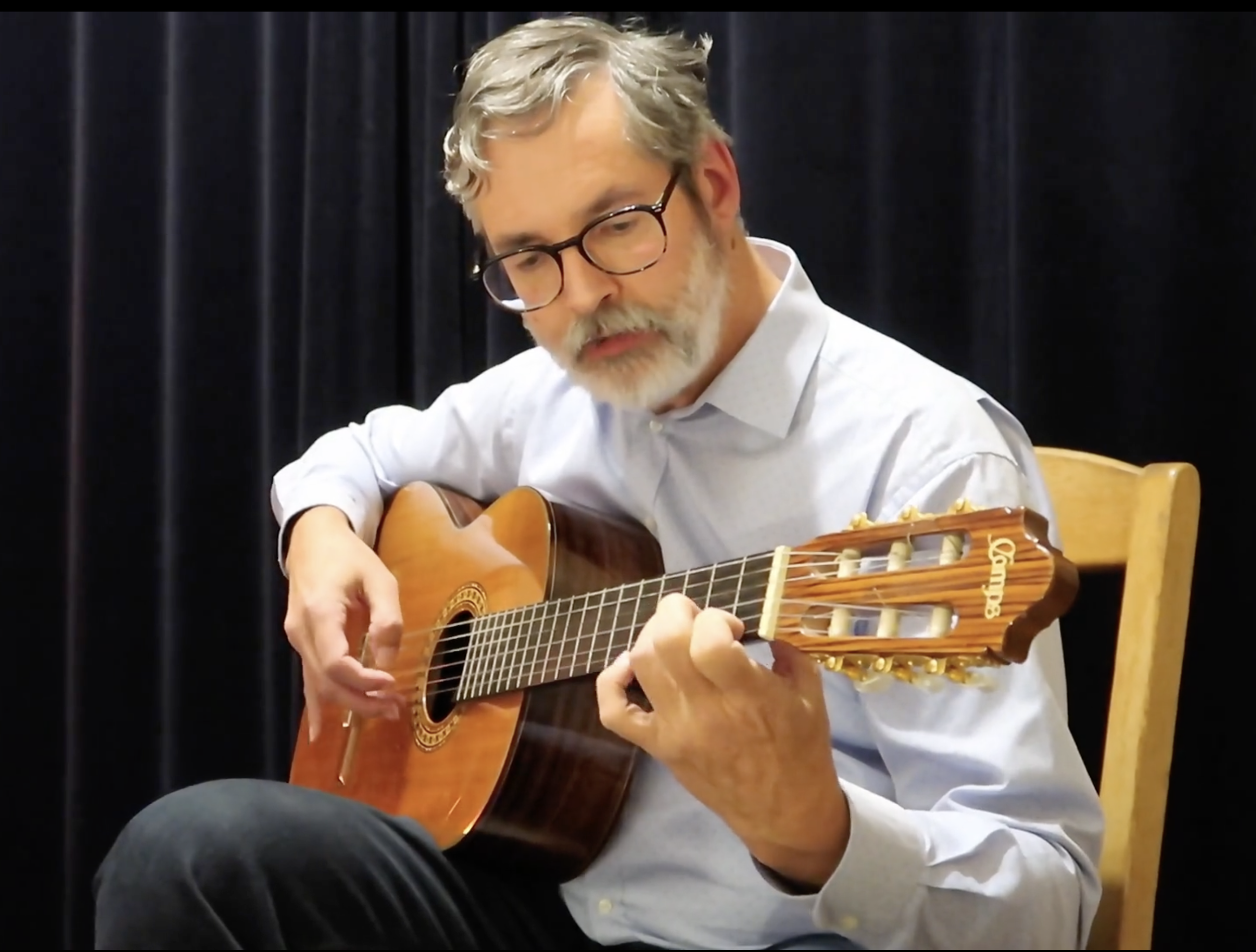 Un homme avec des lunettes jouent de la guitare acoustique devant un fond noir.