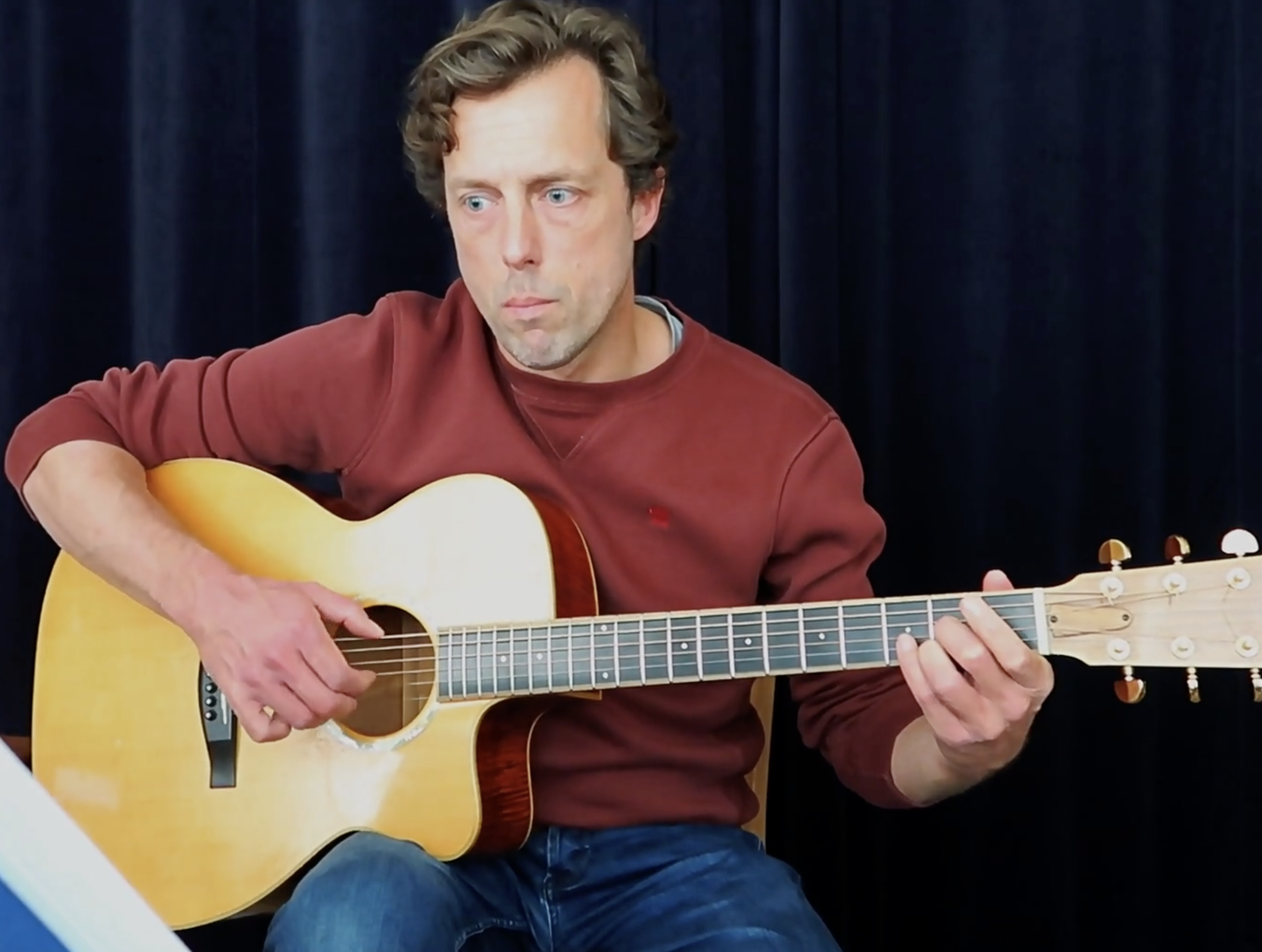 Un homme jouant de la guitare acoustique, assis devant un fond de rideaux noirs, portant un sweat rouge