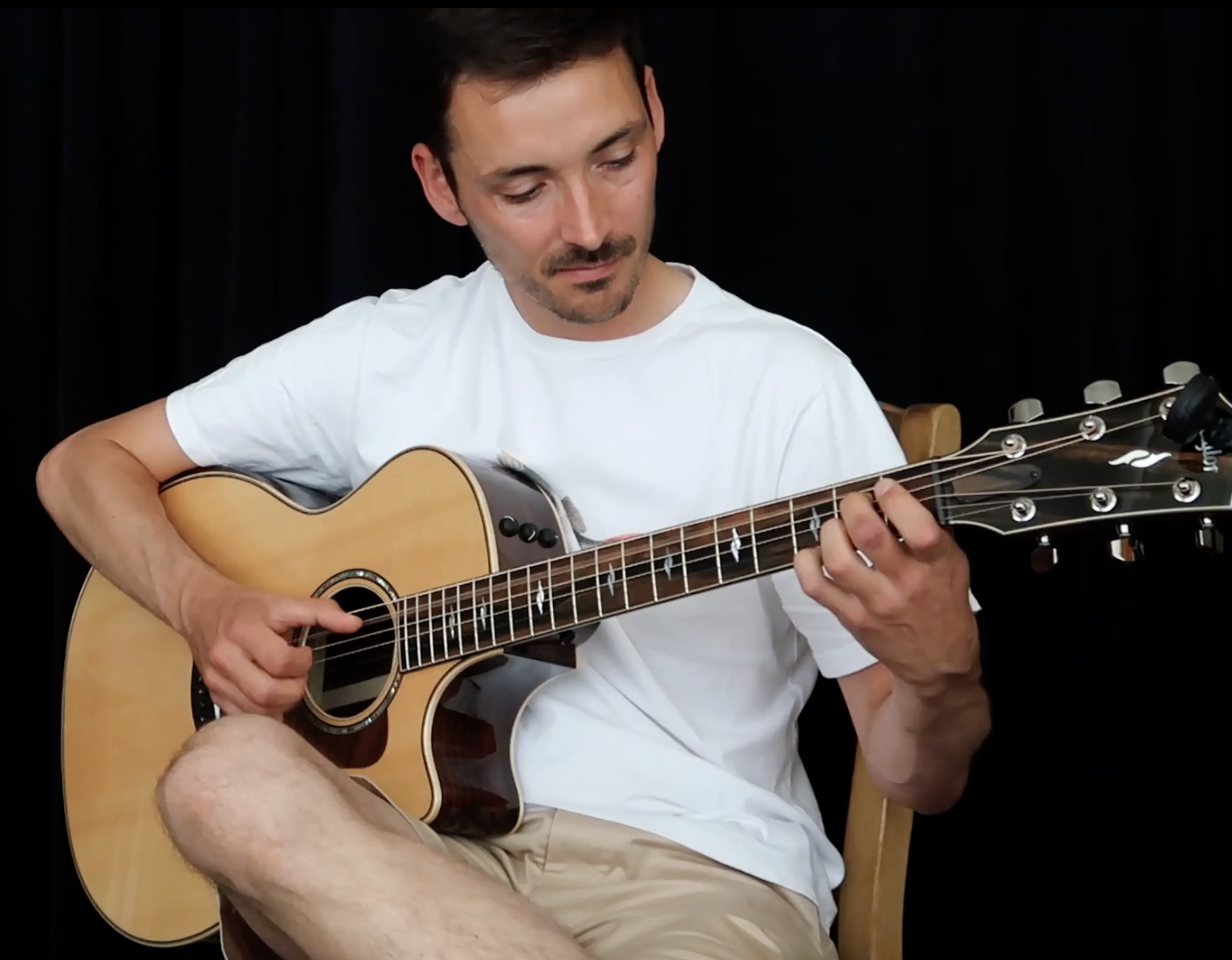 Un homme joue de la guitare acoustique en regardant vers le bas, assis sur une chaise, portant un t-shirt blanc et un short beige, avec un fond noir.