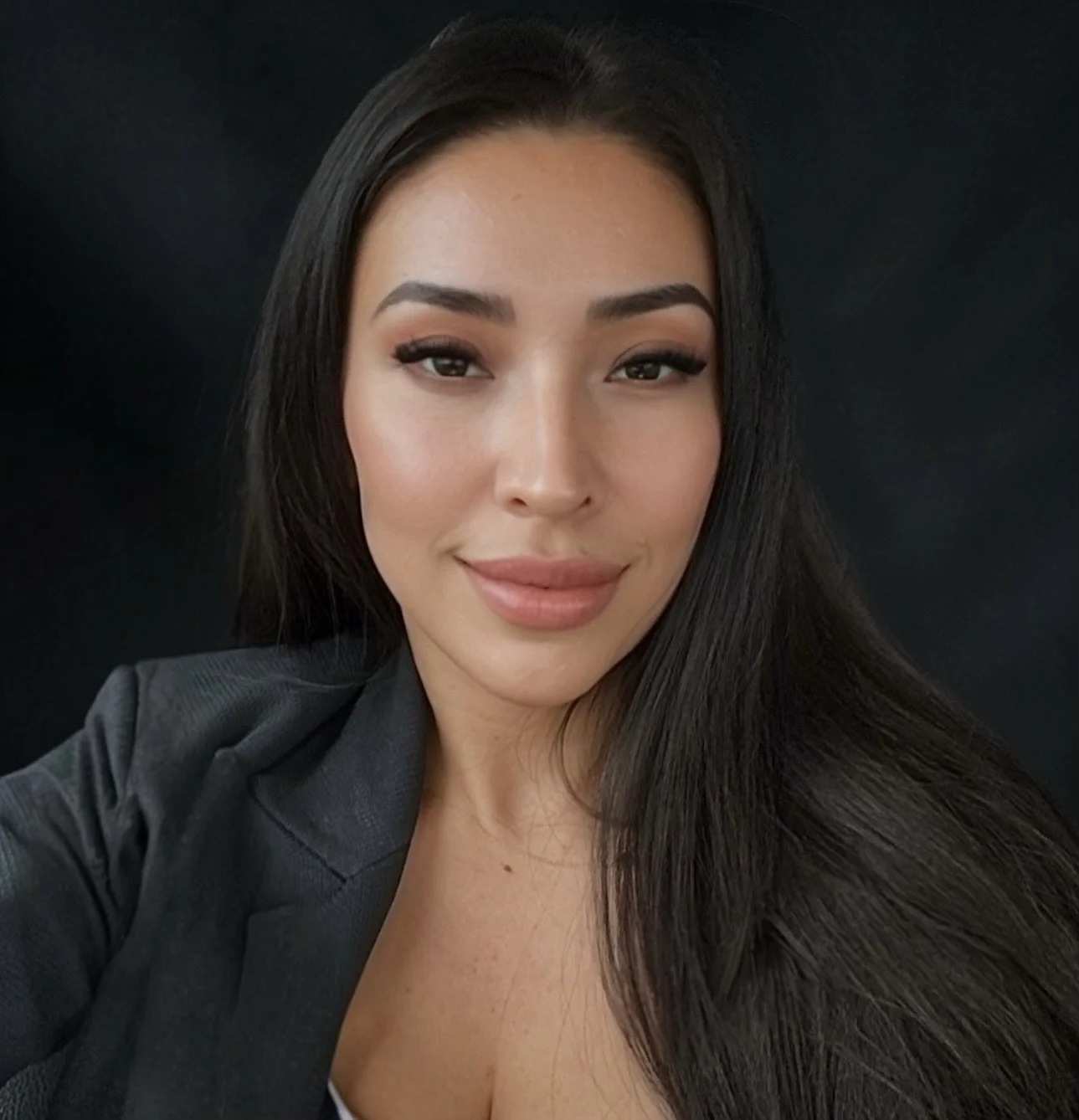 Close-up portrait of a woman with long dark hair, wearing a dark blazer, smiling softly with a dark background.