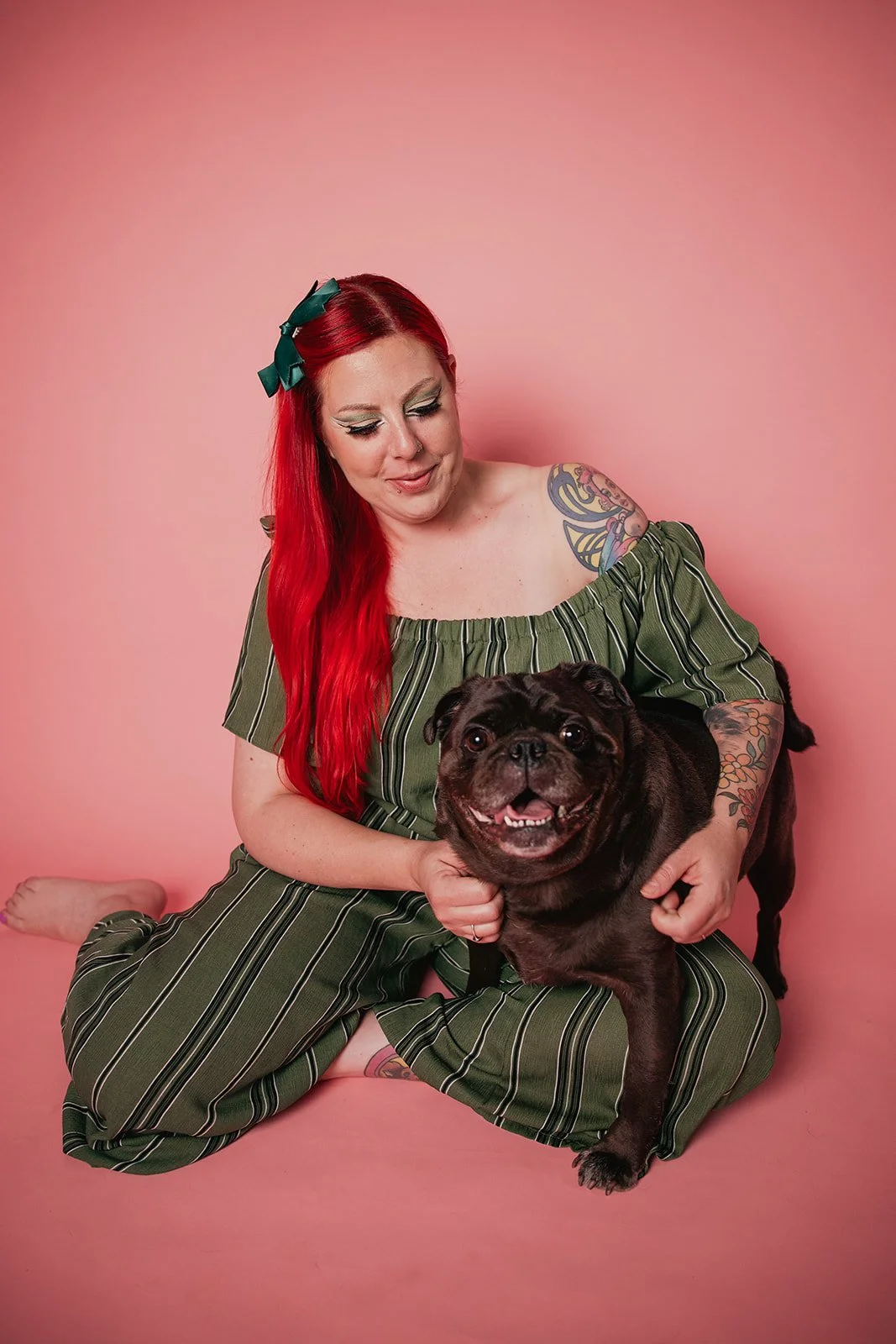 Nadia from Fairy Bread Photo with long red hair, tattoos, and makeup sitting on a pink background, holding a happy black pug.