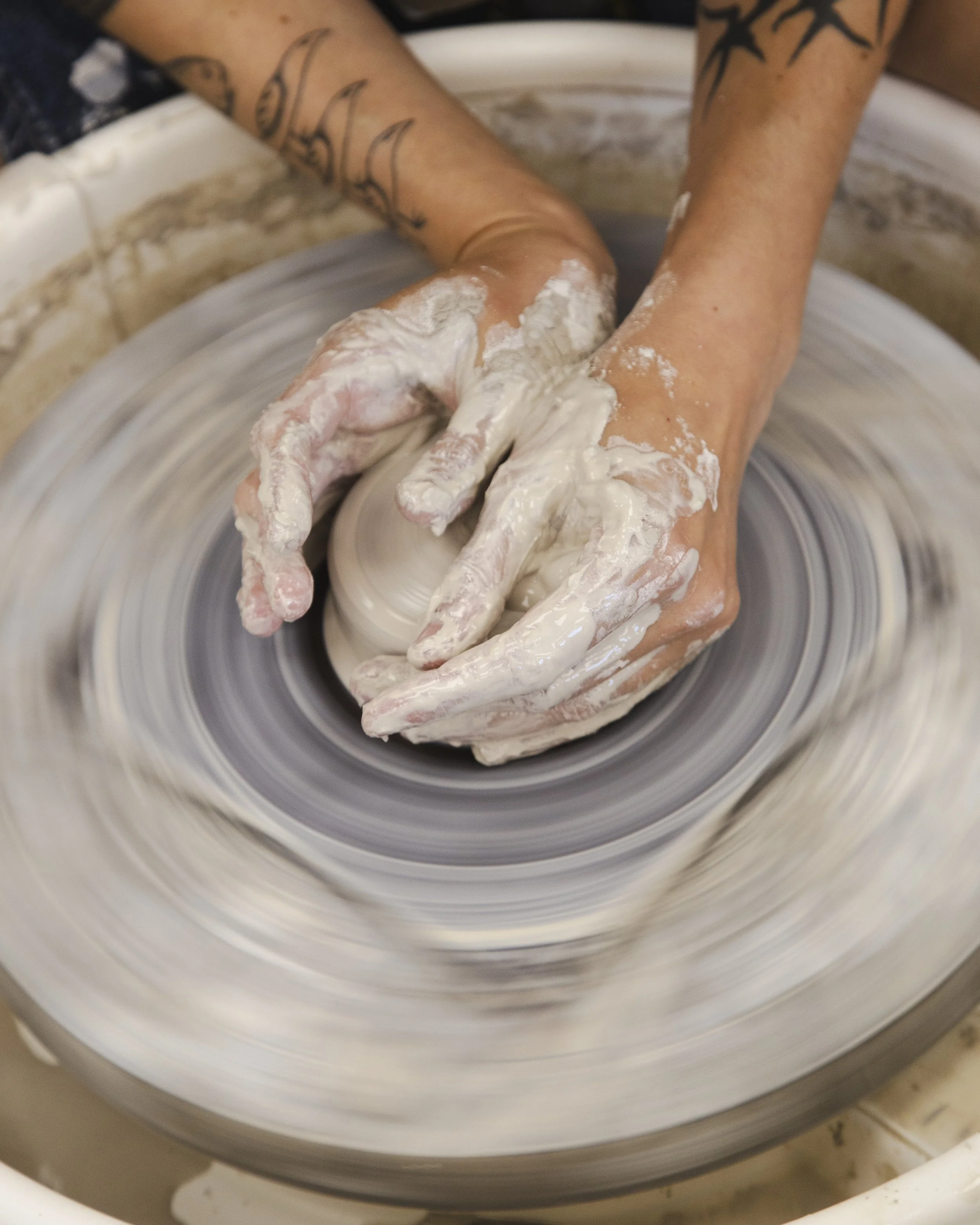 A person’s hands covered in white clay, shaping a pottery piece on a spinning pottery wheel at Still Life Ceramics in Richmond, VA
