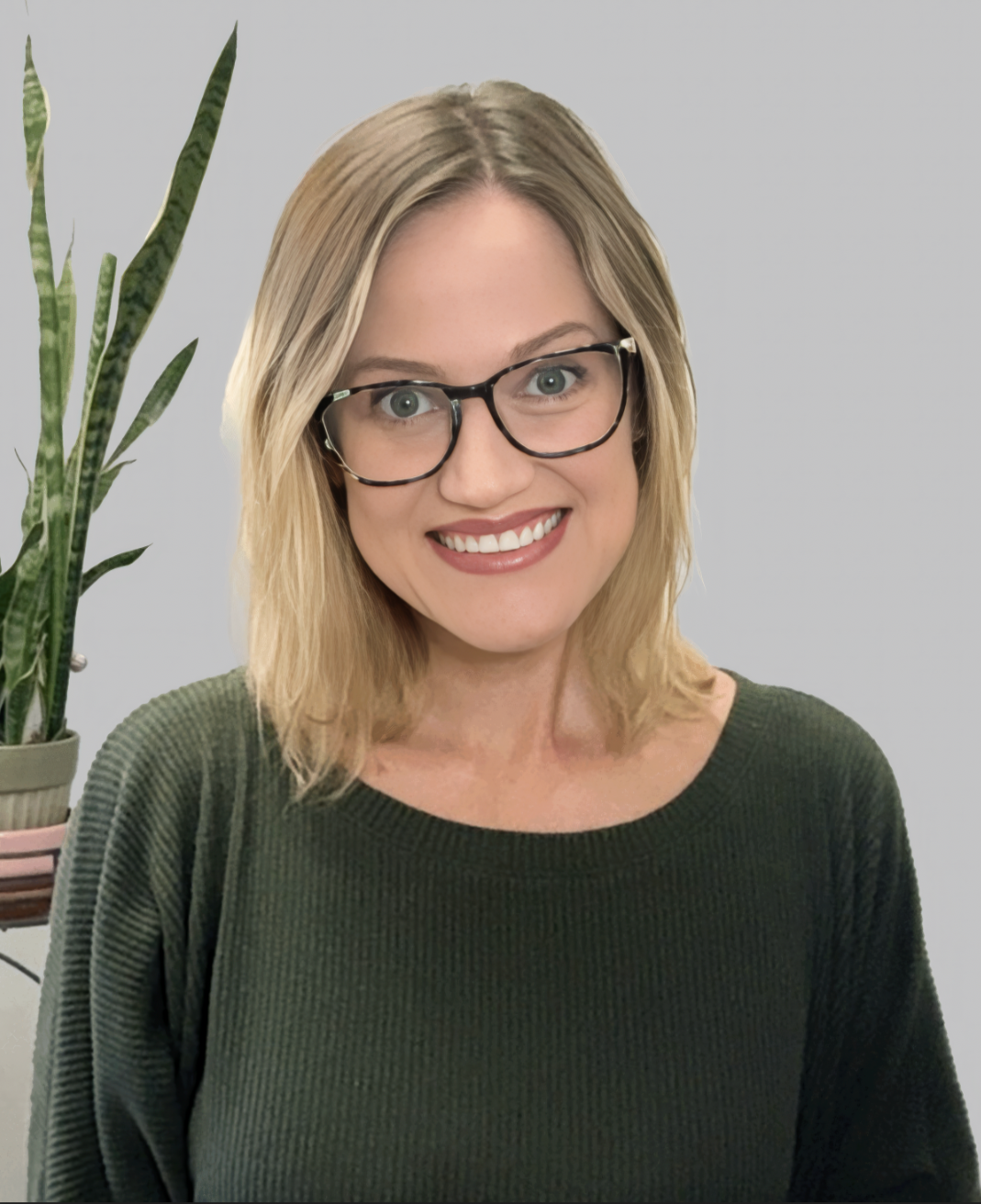 A woman with blonde hair, glasses, and a green sweater smiling at the camera, standing next to a potted plant against a gray background.