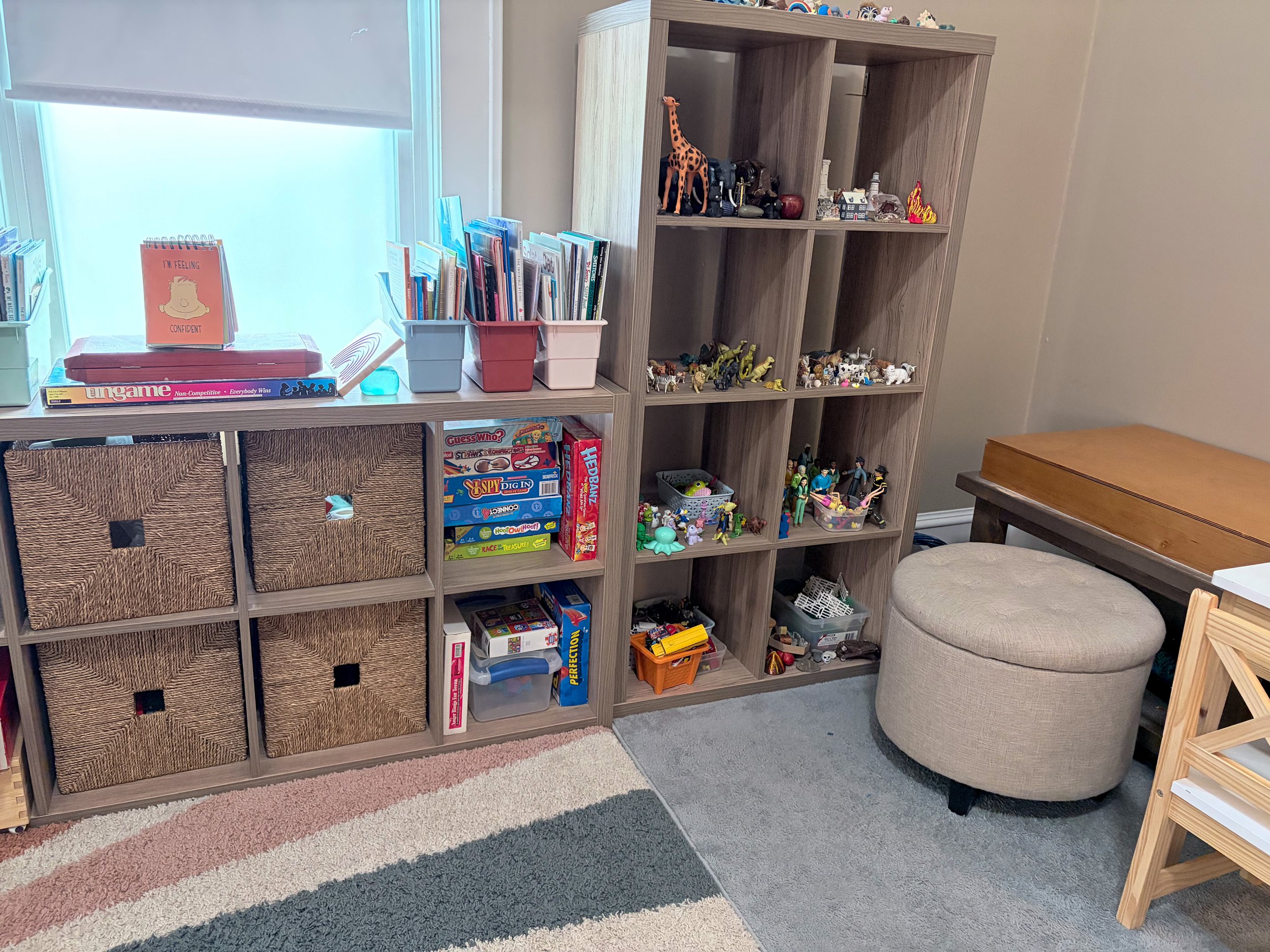 Child's playroom corner with wooden shelves filled with toys, books, and games, tan ottoman, wooden table, storage baskets, and a colorful rug.