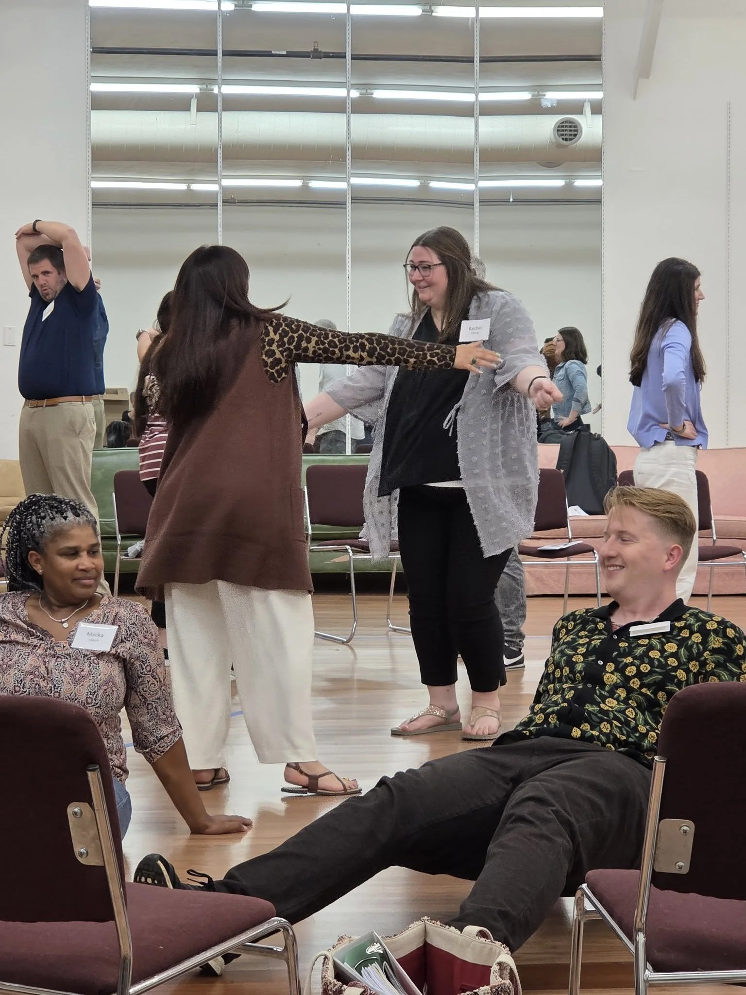 People engage in a social activity in a room with chairs and a large mirror. A woman with glasses and a gray top dances with a woman in a brown top and leopard print sleeves. Others sit or stand around them, smiling and observing.