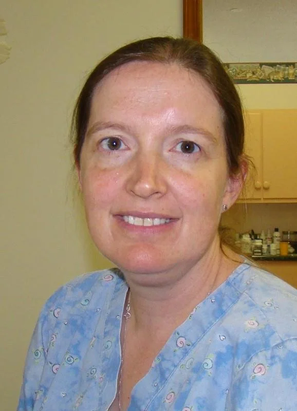 A woman with light skin, reddish-brown hair, and blue eyes, wearing blue medical scrubs, standing in a room with a kitchen in the background.