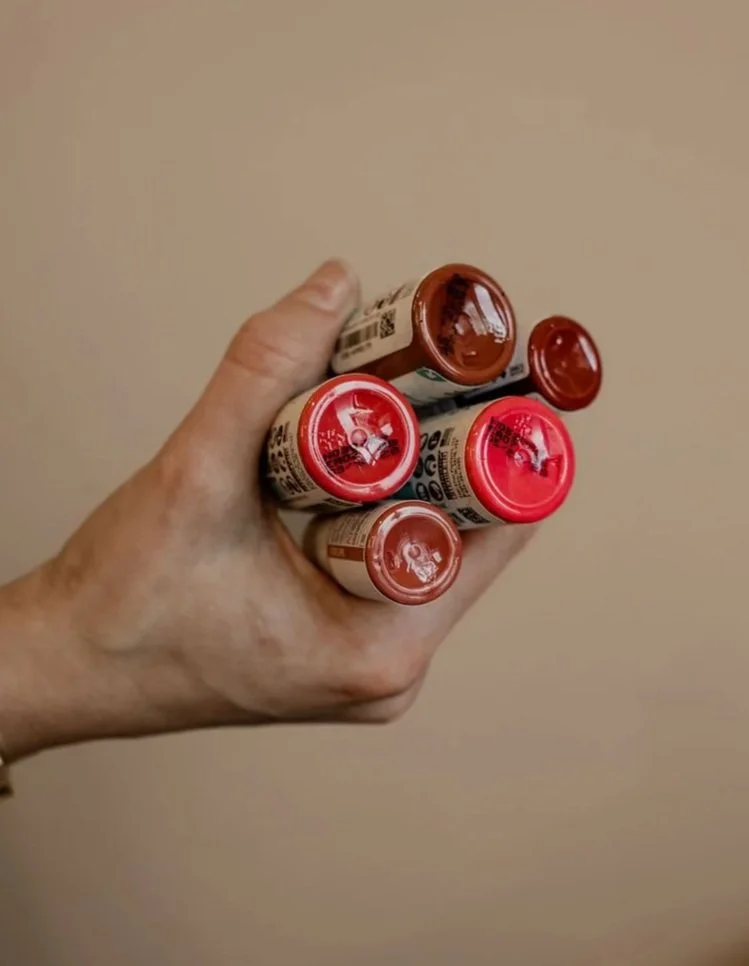 A hand holding five small bottles with different colored caps, likely paint or similar products, against a plain beige background.