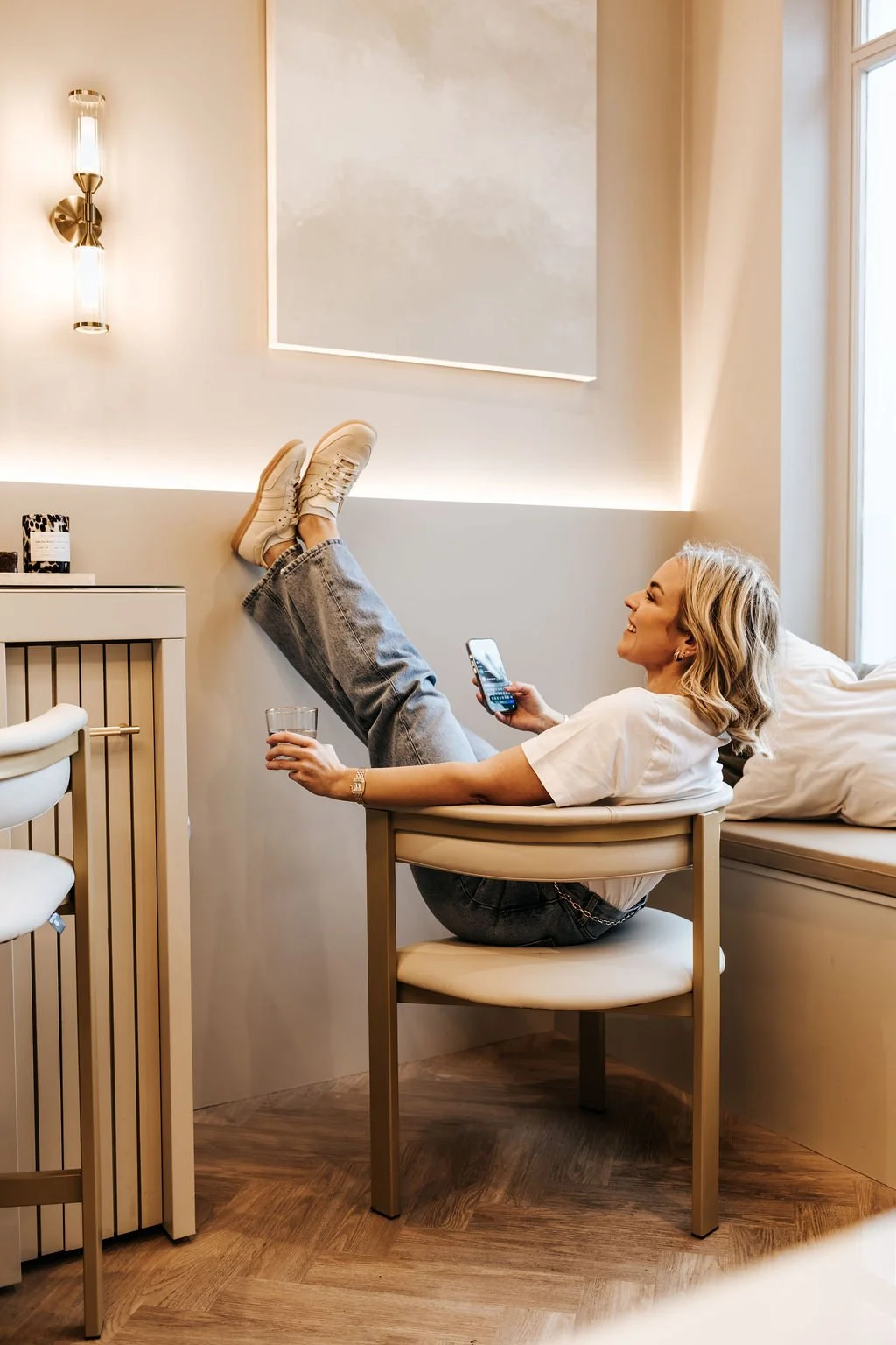 A woman relaxing in a modern, bright room, sitting in a beige chair with her feet propped up on a white cabinet. She is smiling, holding a glass in one hand and a phone in the other, wearing casual white top, blue jeans, and white sneakers. There is a window letting in natural light and a minimalistic decor style.