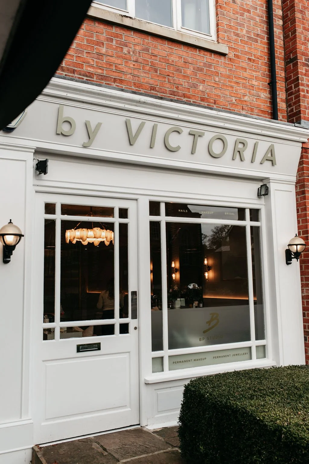 Exterior of a shop named 'by VICTORIA' with a white facade, glass windows, and doors, and brick walls. There are black wall-mounted lamps on either side of the entrance, and the shop offers services like permanent makeup, nails, bridal makeup, and more.