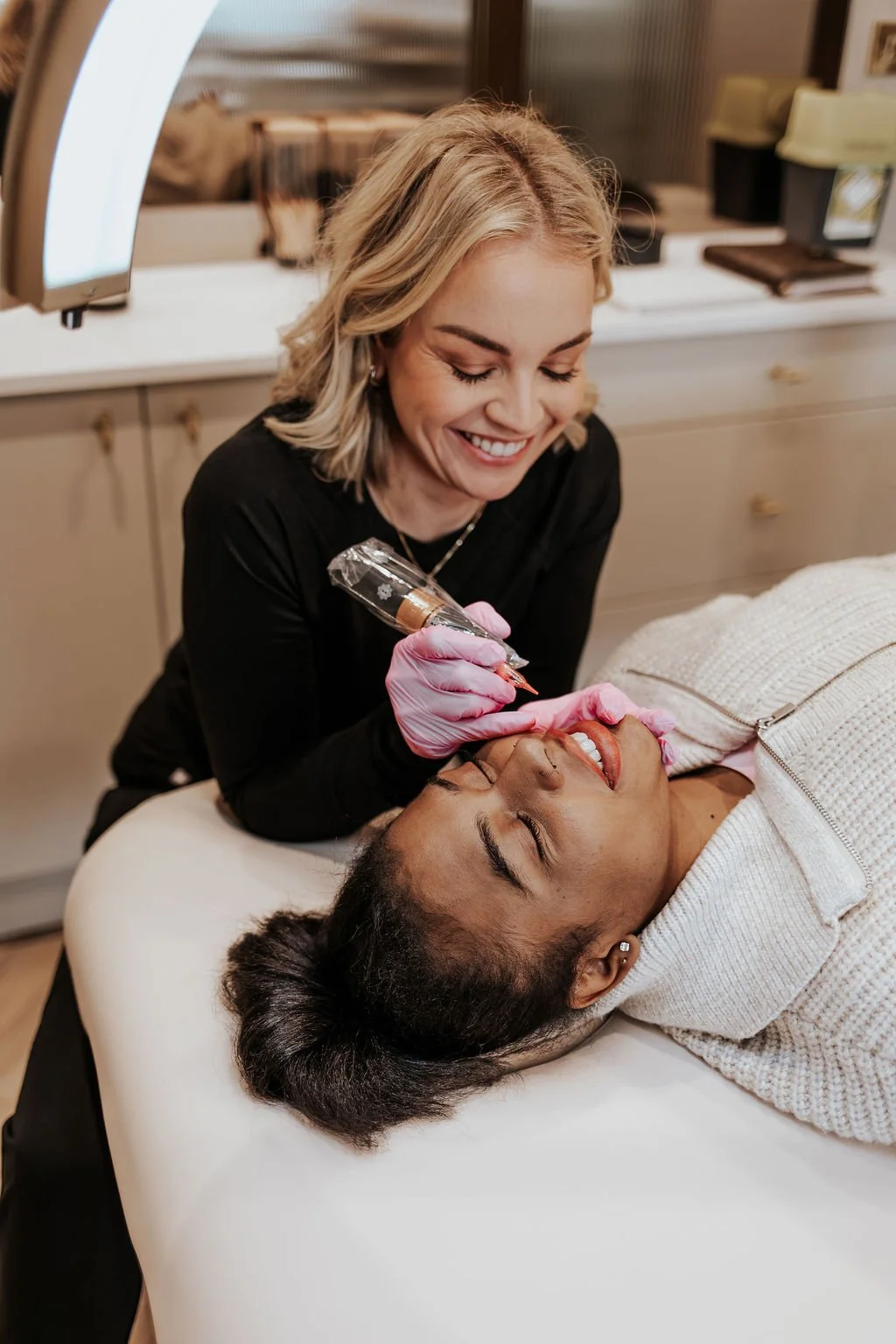 A woman is lying on a bed receiving a cosmetic procedure, while another woman with blonde hair, wearing pink gloves, uses a pen-like device near her lip. The setting appears to be a clinic or spa.