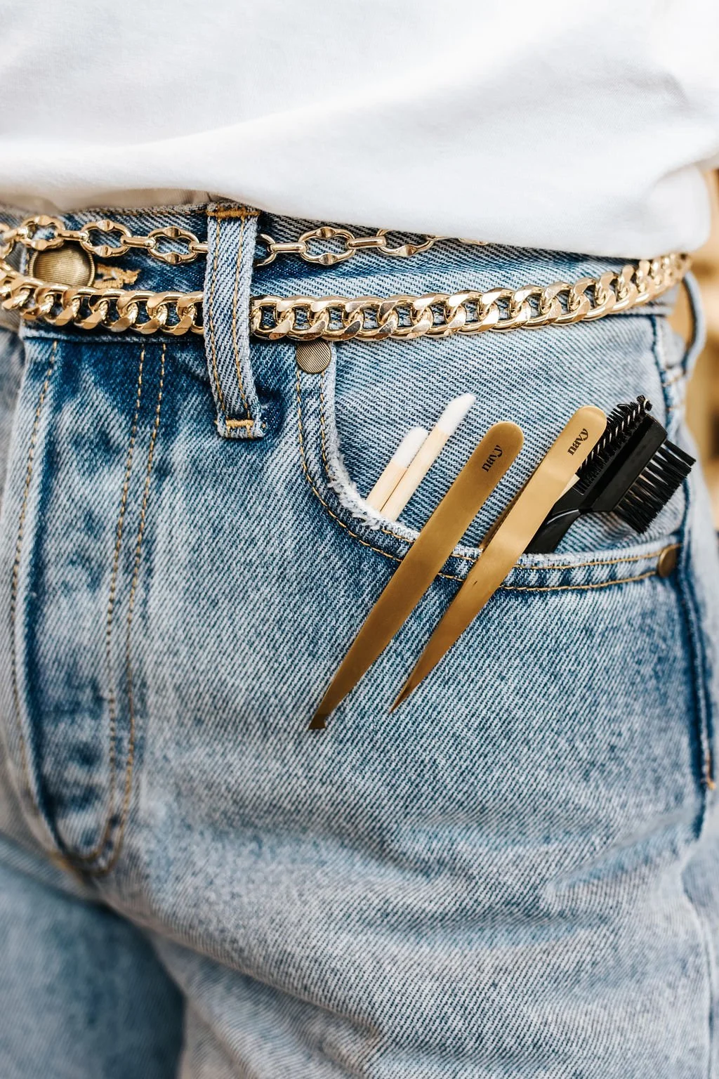 Close-up of a person's jeans pocket with beauty tools including combs, tweezers, and a small brush, and a white cotton swab, with a white shirt and gold chain belt.