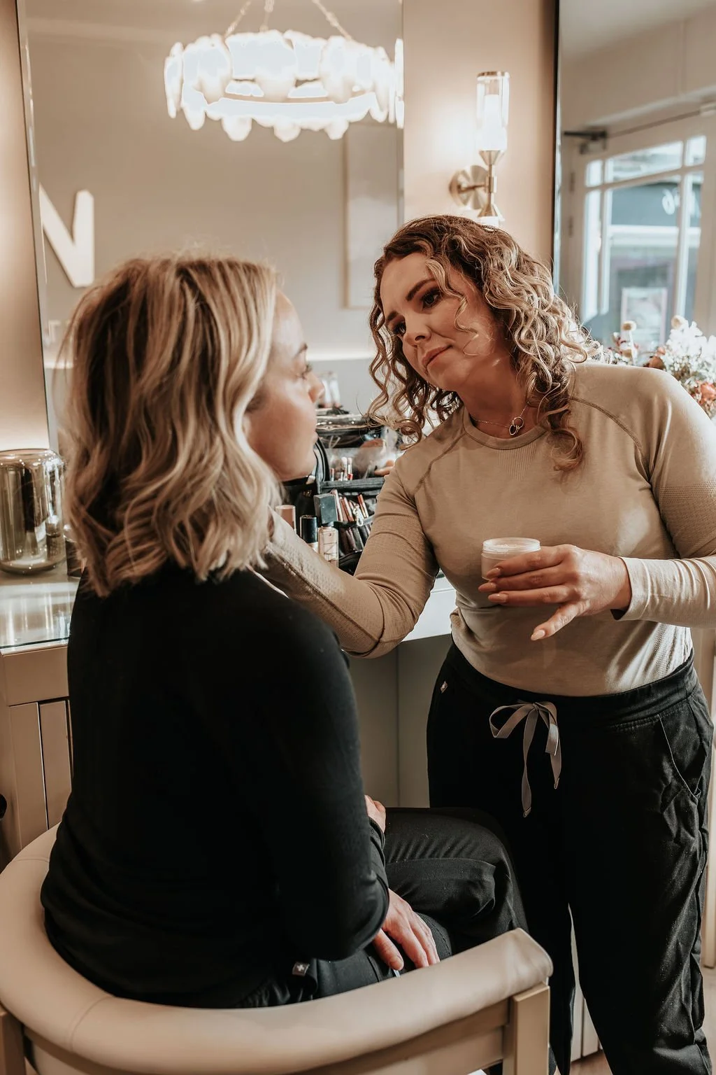A makeup artist is applying makeup to a woman seated in a salon, with a mirror and makeup products nearby.
