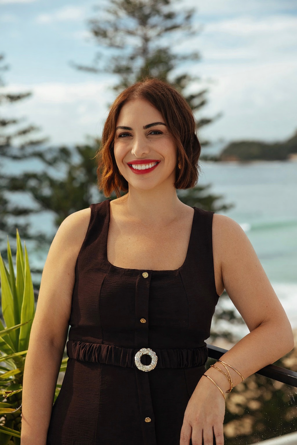 A woman with short brown hair and red lipstick smiling outdoors with a background of trees and water.