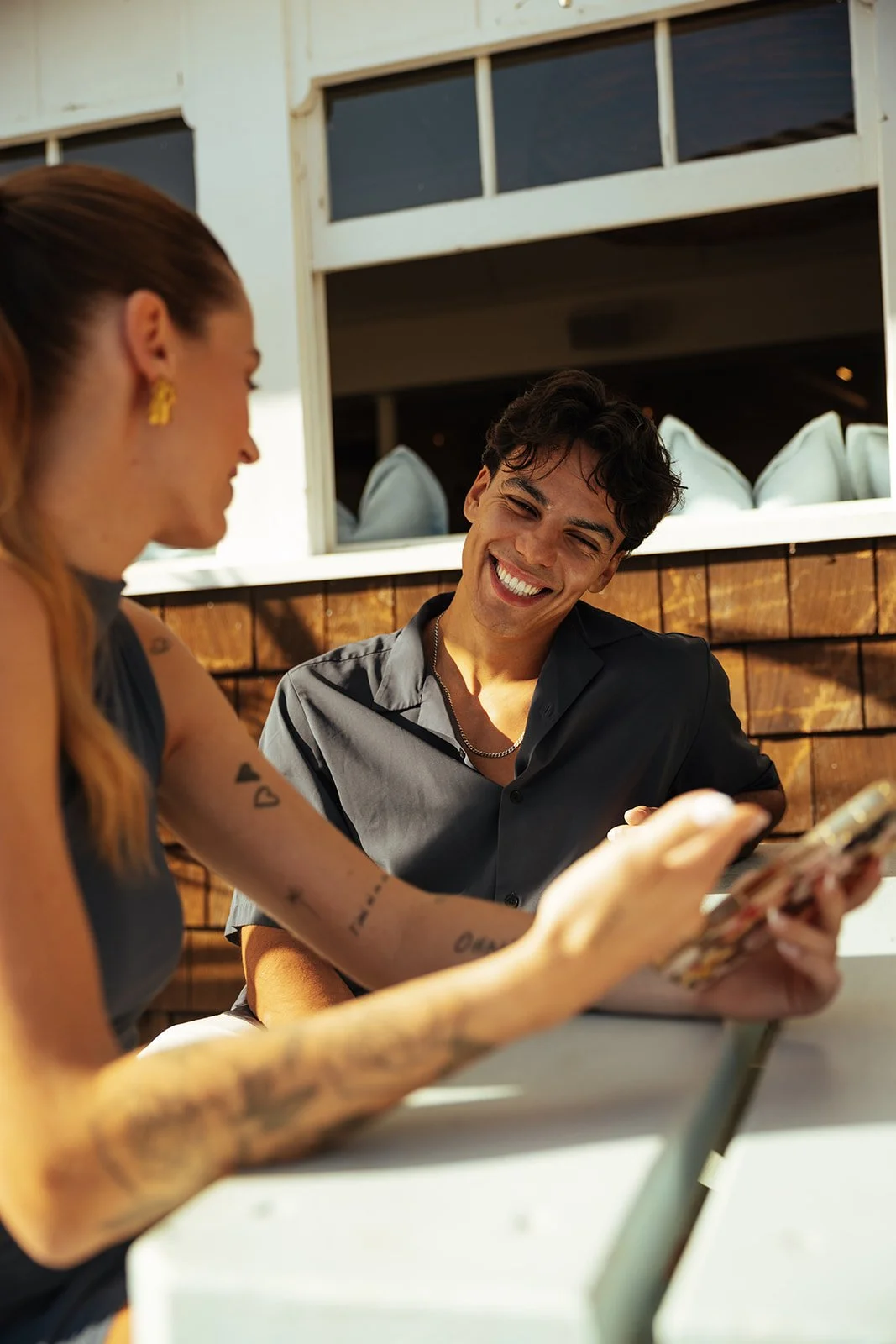 A man and woman sitting at an outdoor table, laughing and looking at a phone.