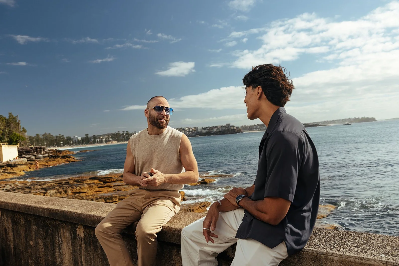 Two men are sitting and talking by the water on a sunny day, with a beach and buildings in the background.