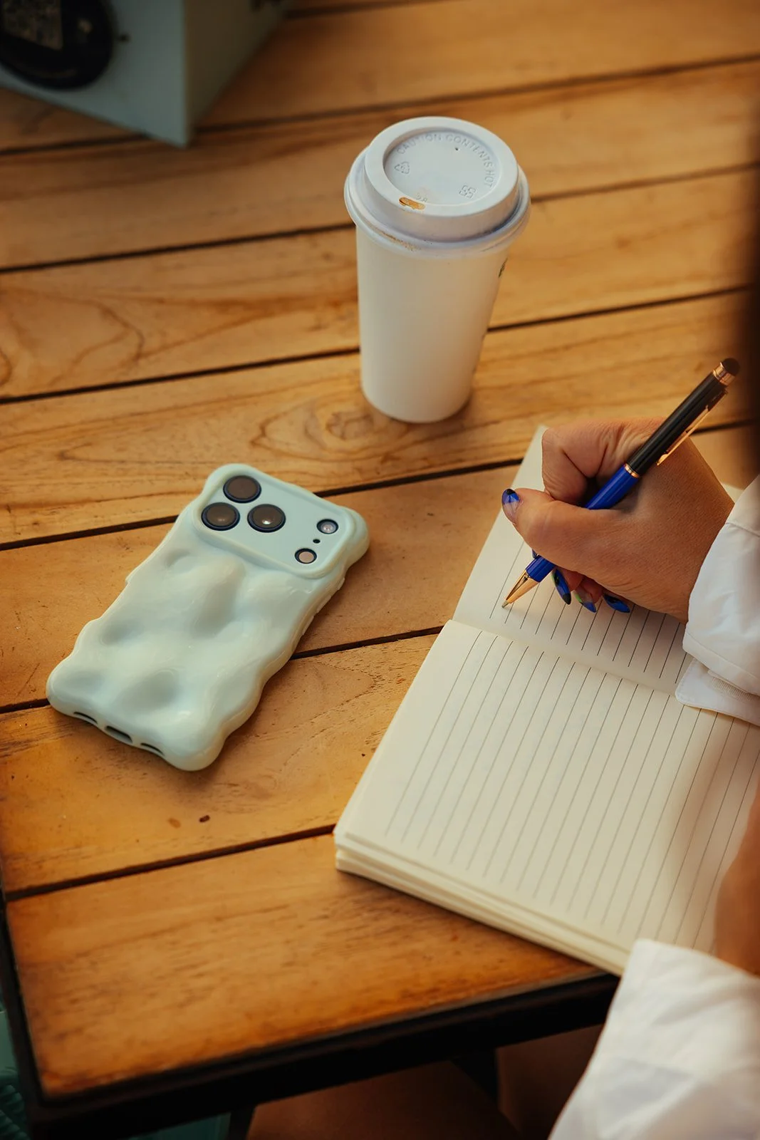 A person writing in a lined notebook with a blue pen on a wooden table. Nearby are a white tech device with a mint green case resembling a Tetris block, and a white disposable coffee cup with a lid.