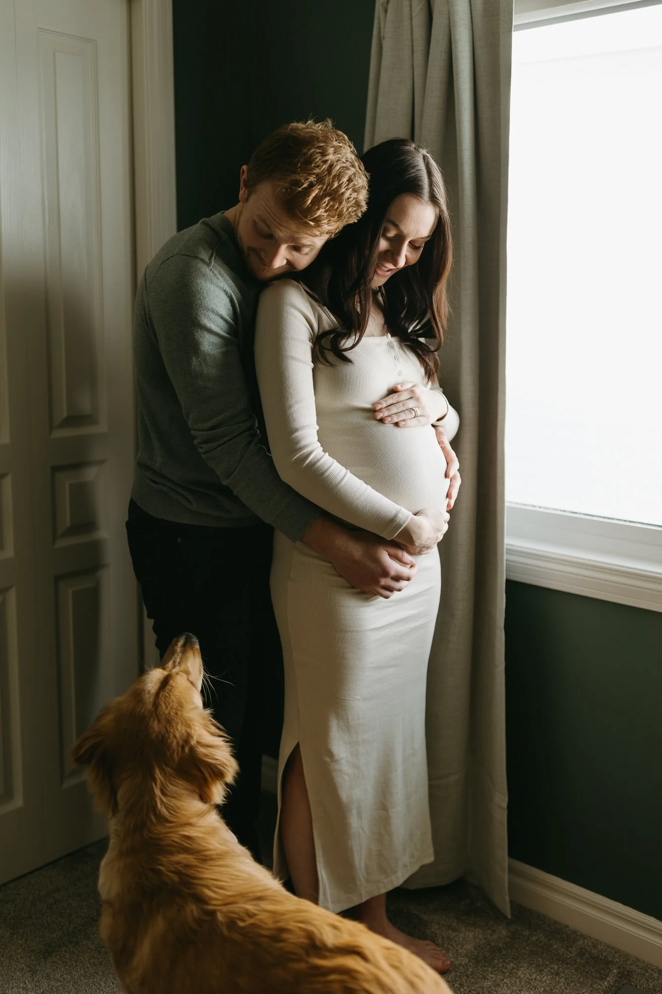 A couple with a pregnant woman standing close together near a window, with the man hugging her and a dog looking up at them.