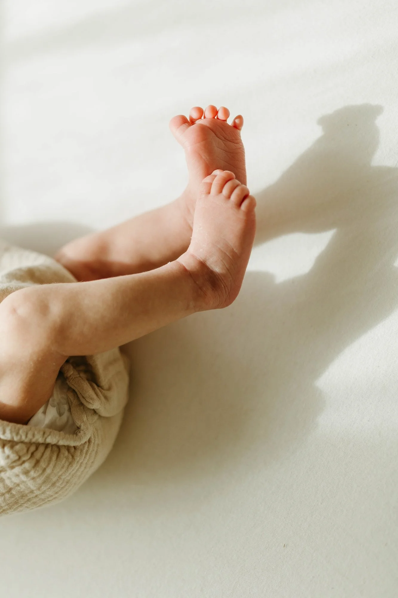 Close-up of a baby's feet with adult fingers gently holding one foot, casting a shadow on a white surface.