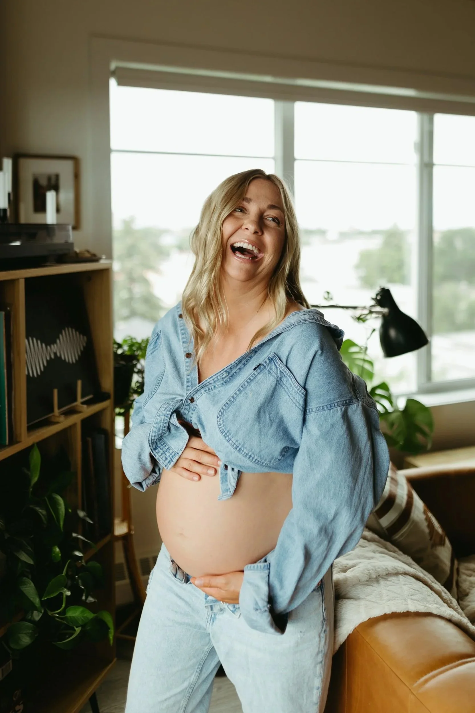 A pregnant woman laughing while holding her belly in a cozy living room with large windows, a plant, and a bookshelf.