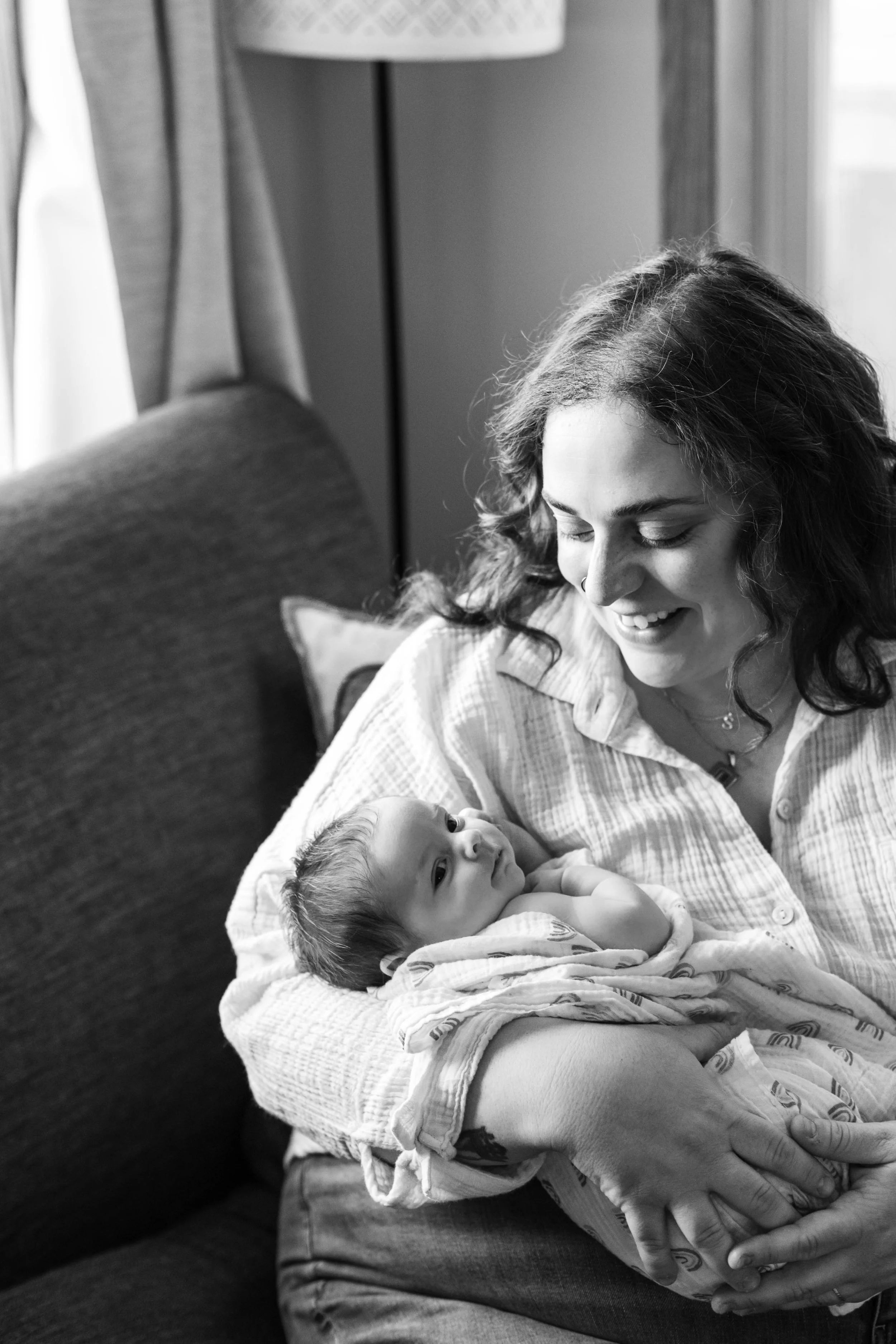 A woman sitting on the couch near a window gazes down sweetly at her newborn baby, wrapped in a blanket 