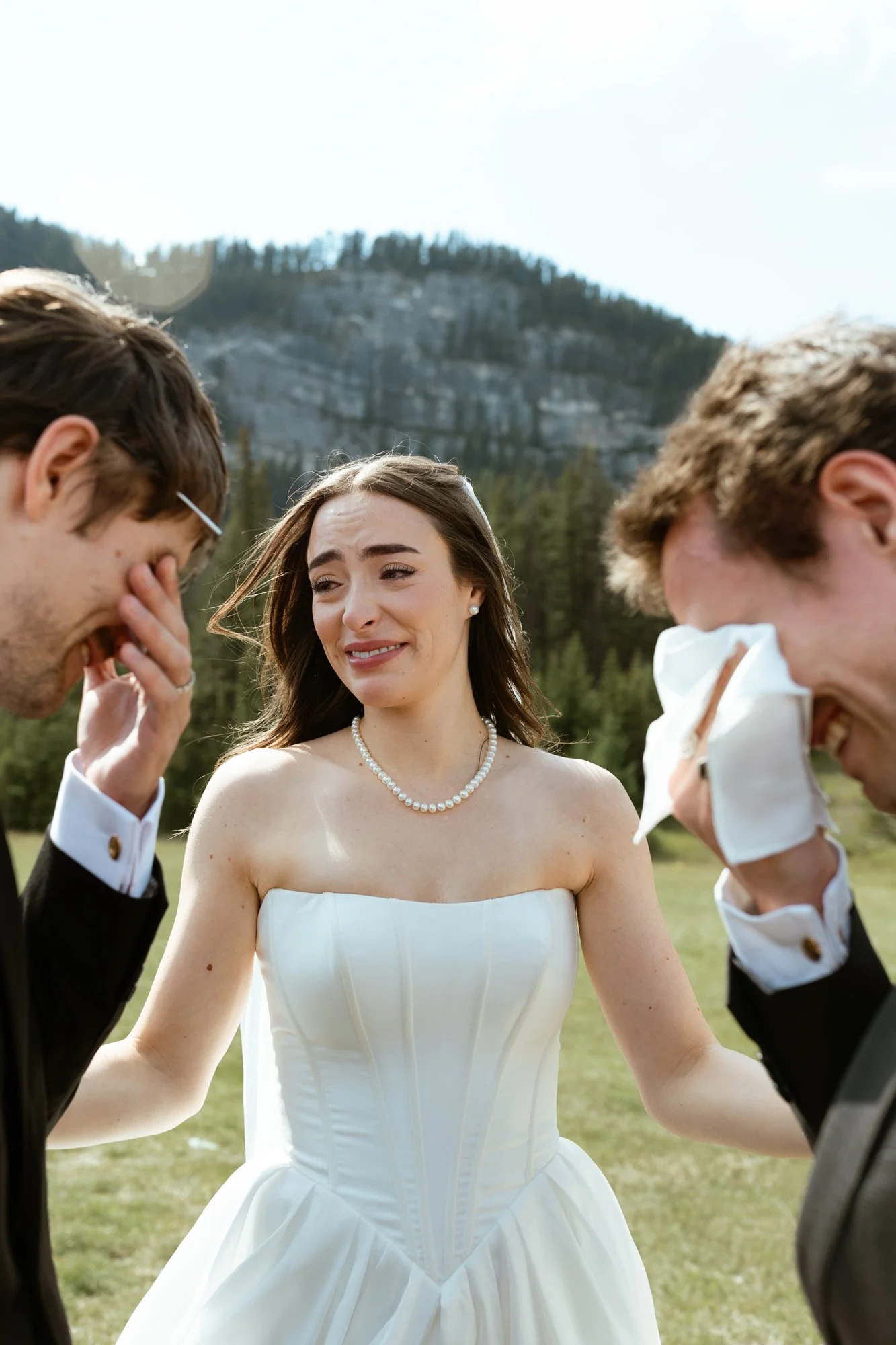 A woman in a wedding dress with a pearl necklace is emotional, standing outdoors with two men who are also emotional, possibly during a wedding ceremony in a natural landscape with mountains and trees.