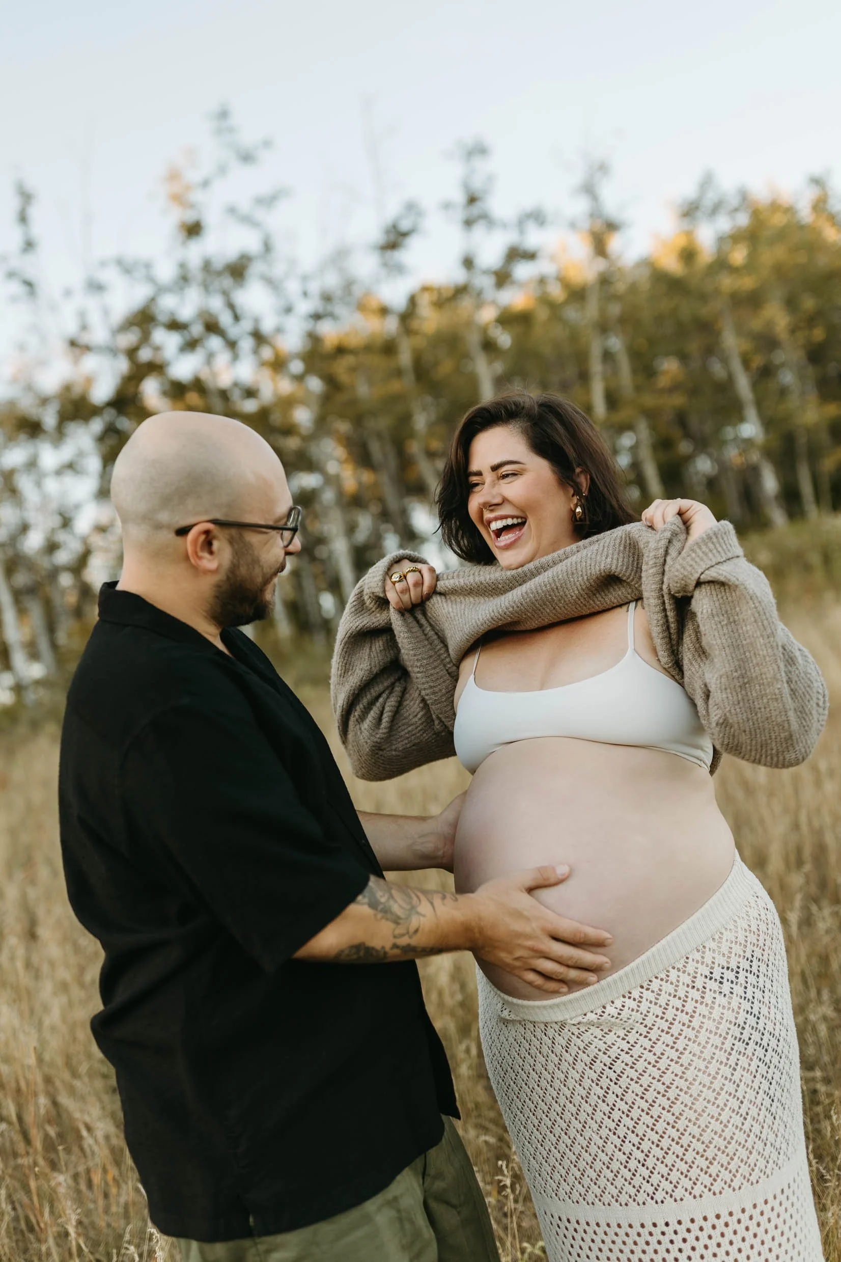 Candid photo of pregnant woman laughing as she lifts up her sweater to show her husband her belly at her fall maternity session in Calgary