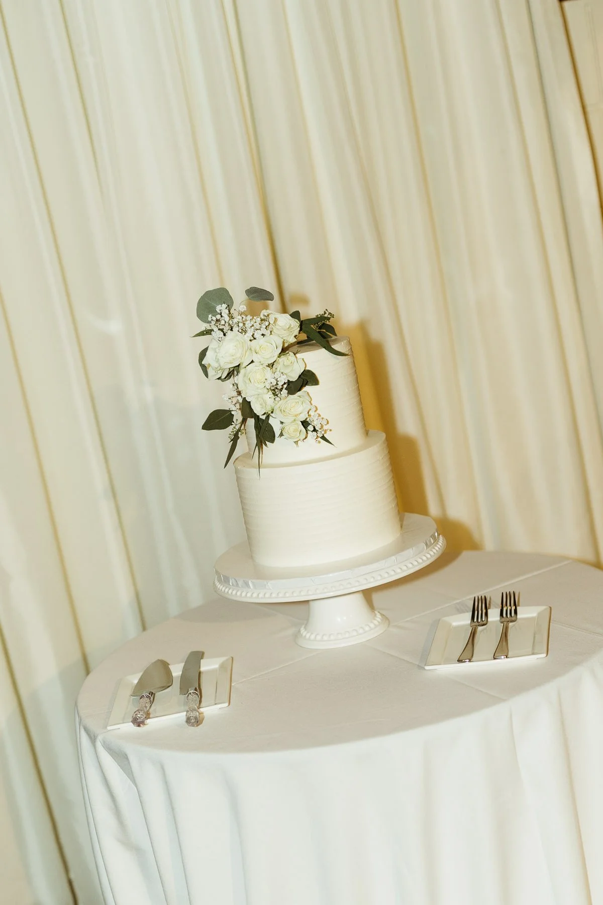 Two-tier white wedding cake with floral decoration on a round table, next to a cake knife and serving set.