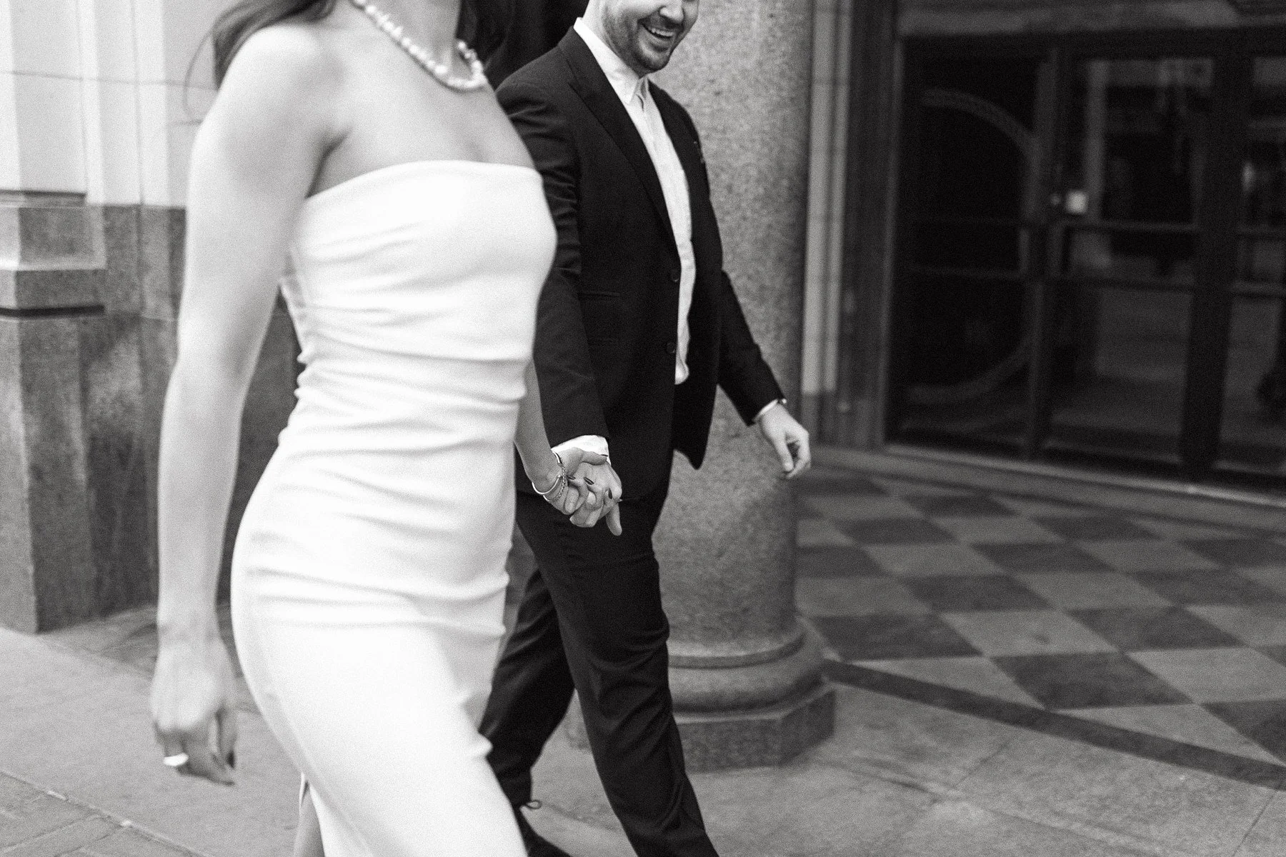 Black and white photo of a couple holding hands, walking in formal attire.