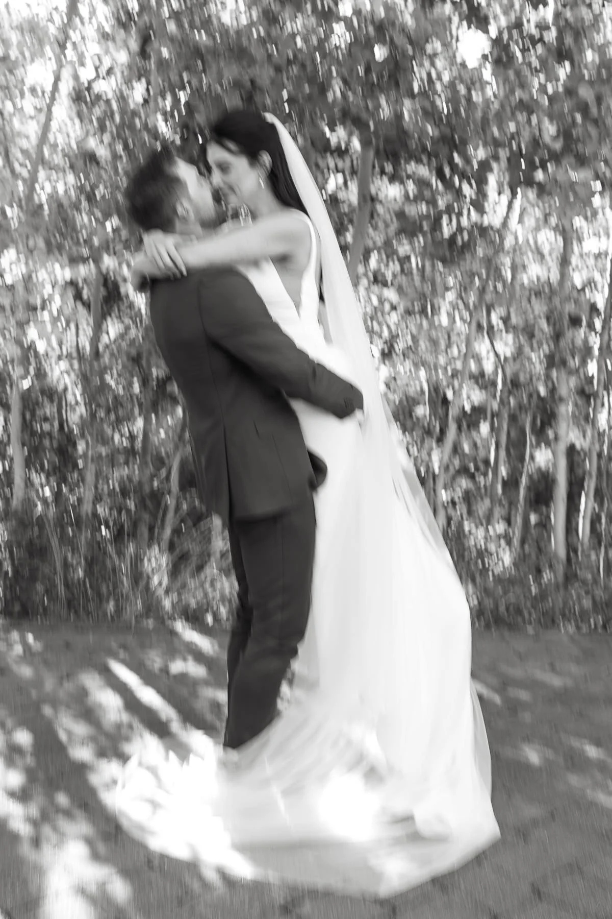 Black and white photo of a groom lifting a bride outdoors, surrounded by trees
