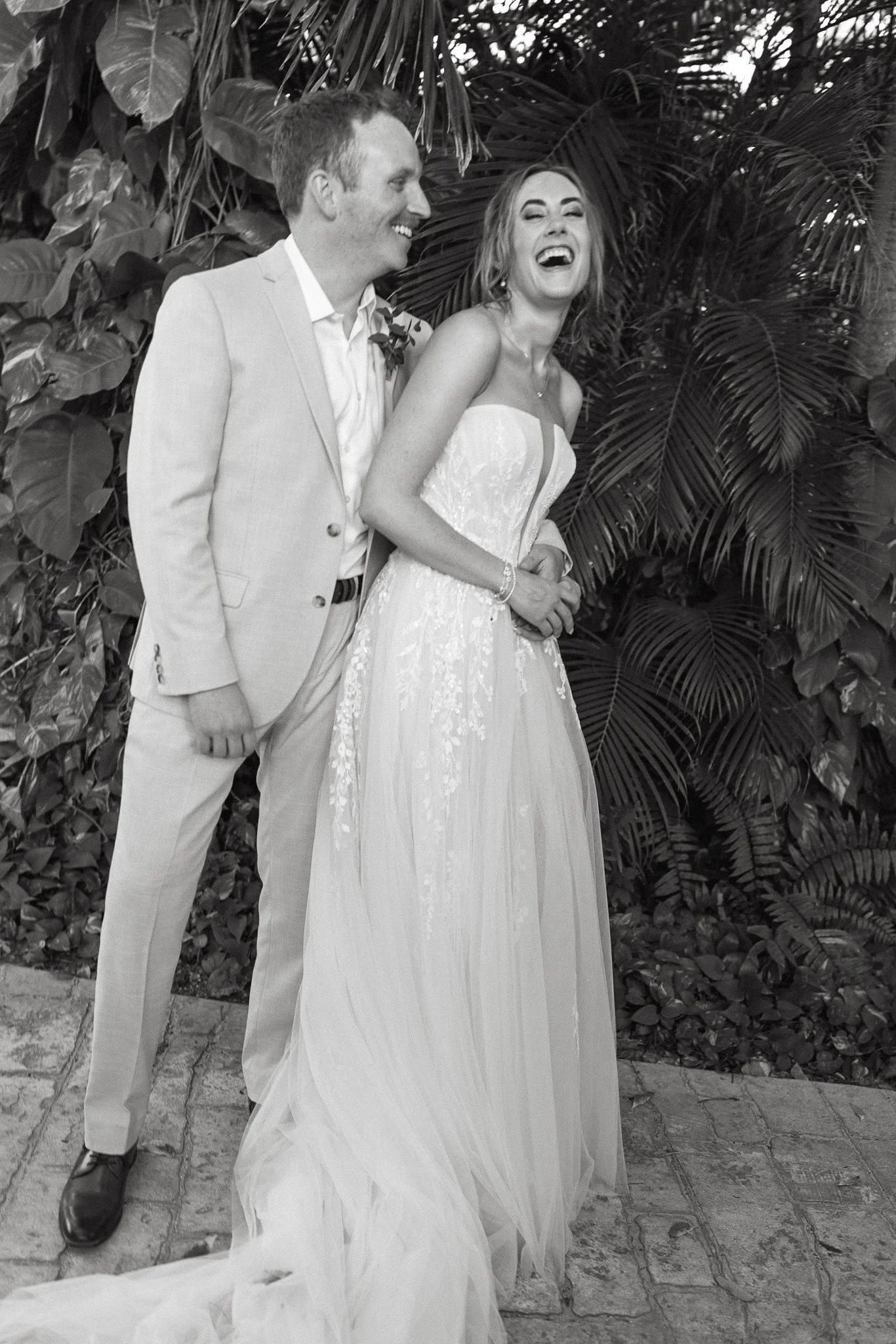 A black and white photo of a bride and groom laughing, standing outdoors in front of lush tropical foliage at The Fives Beach Resort in Playa Del Carmen, Mexico. The groom wears a light-colored suit, and the bride wears a strapless wedding gown .