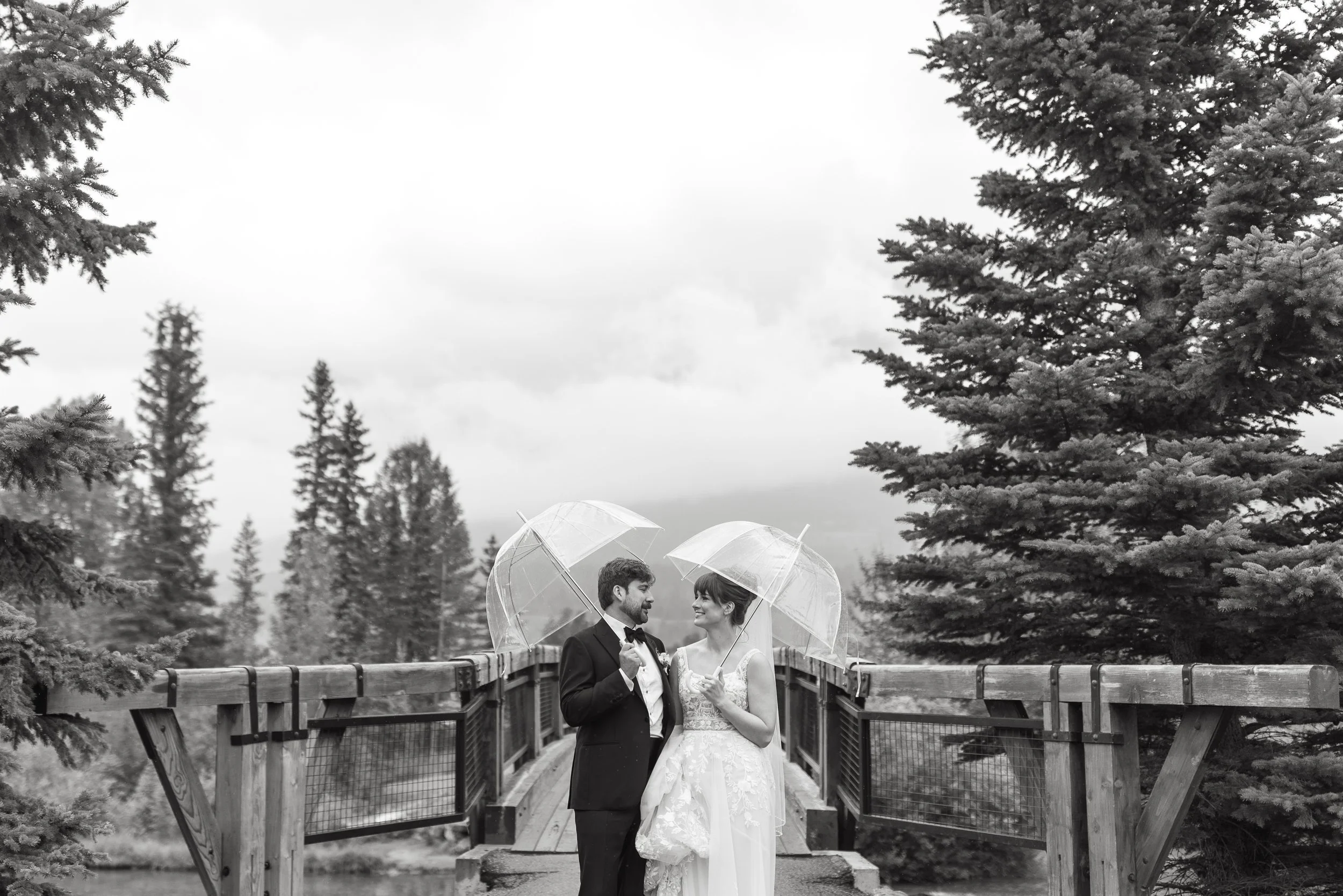 Black and white photo of bride and groom with umbrellas on rainy wedding day in Canmore, Alberta