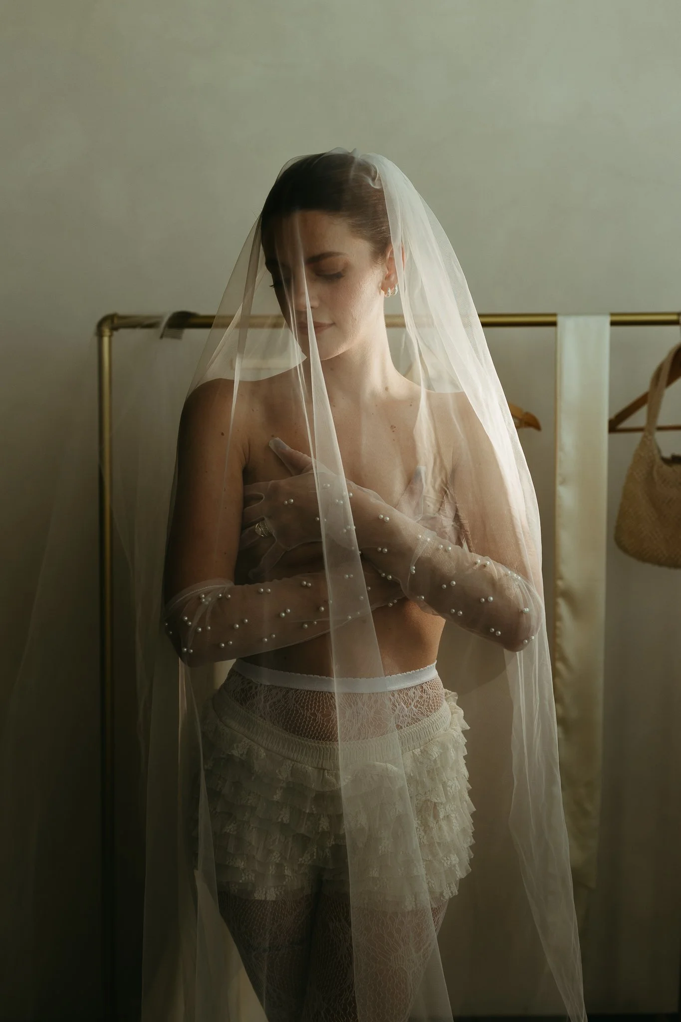 Woman in lace shorts and long veil facing camera, covering chest with arms, wearing pearl-detailed gloves.