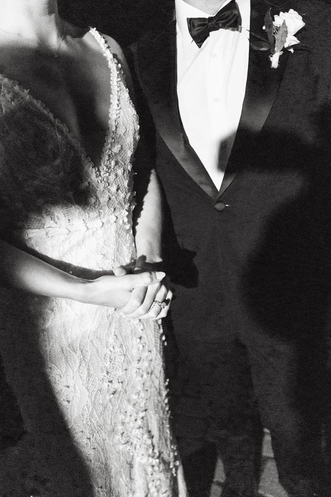 Black and white close-up of a bride and groom holding hands, with the bride wearing a lace wedding dress and the groom in a tuxedo with a bow tie and boutonniere.