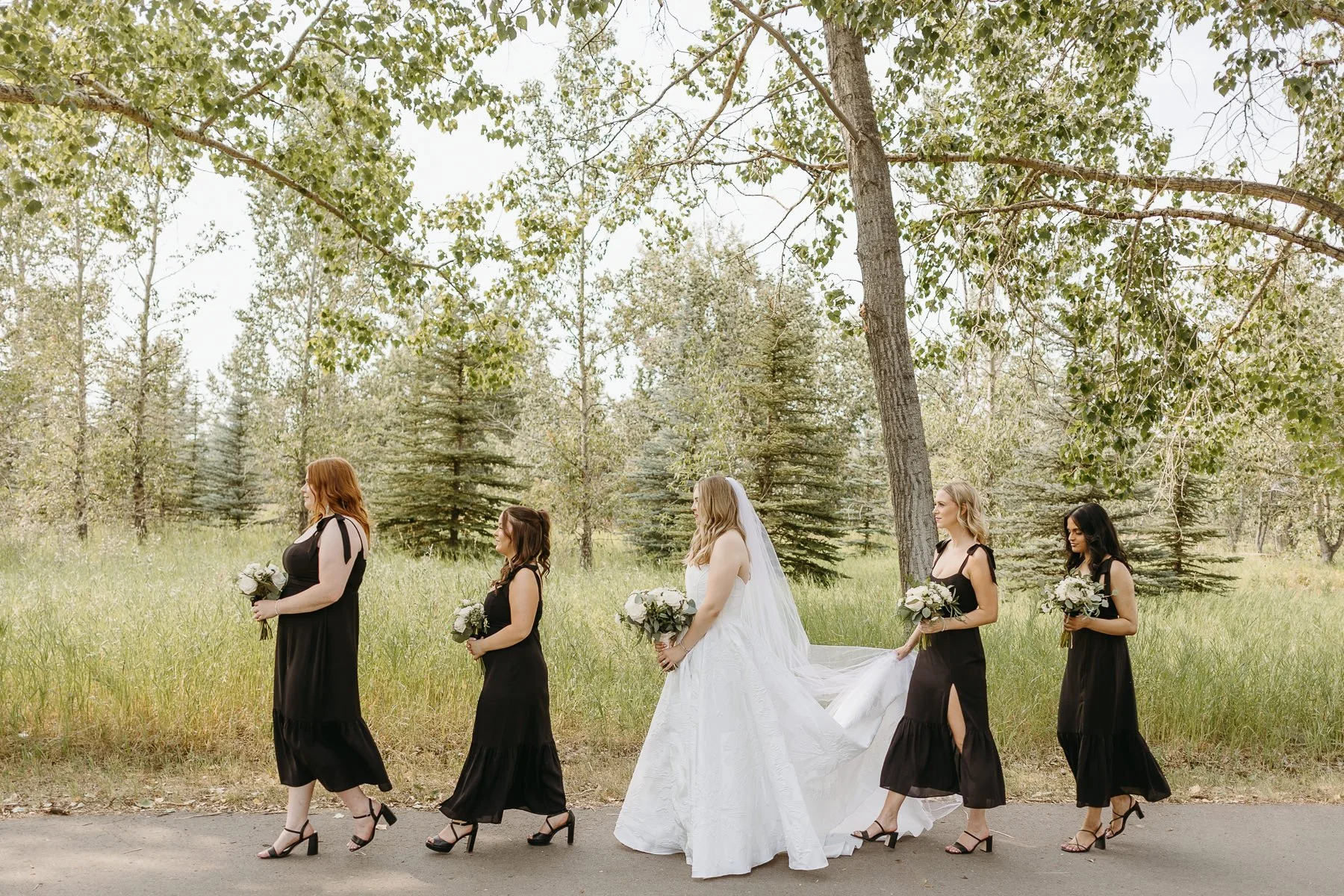 Bridesmaids in black dresses walking with a bride in a white gown outdoors.
