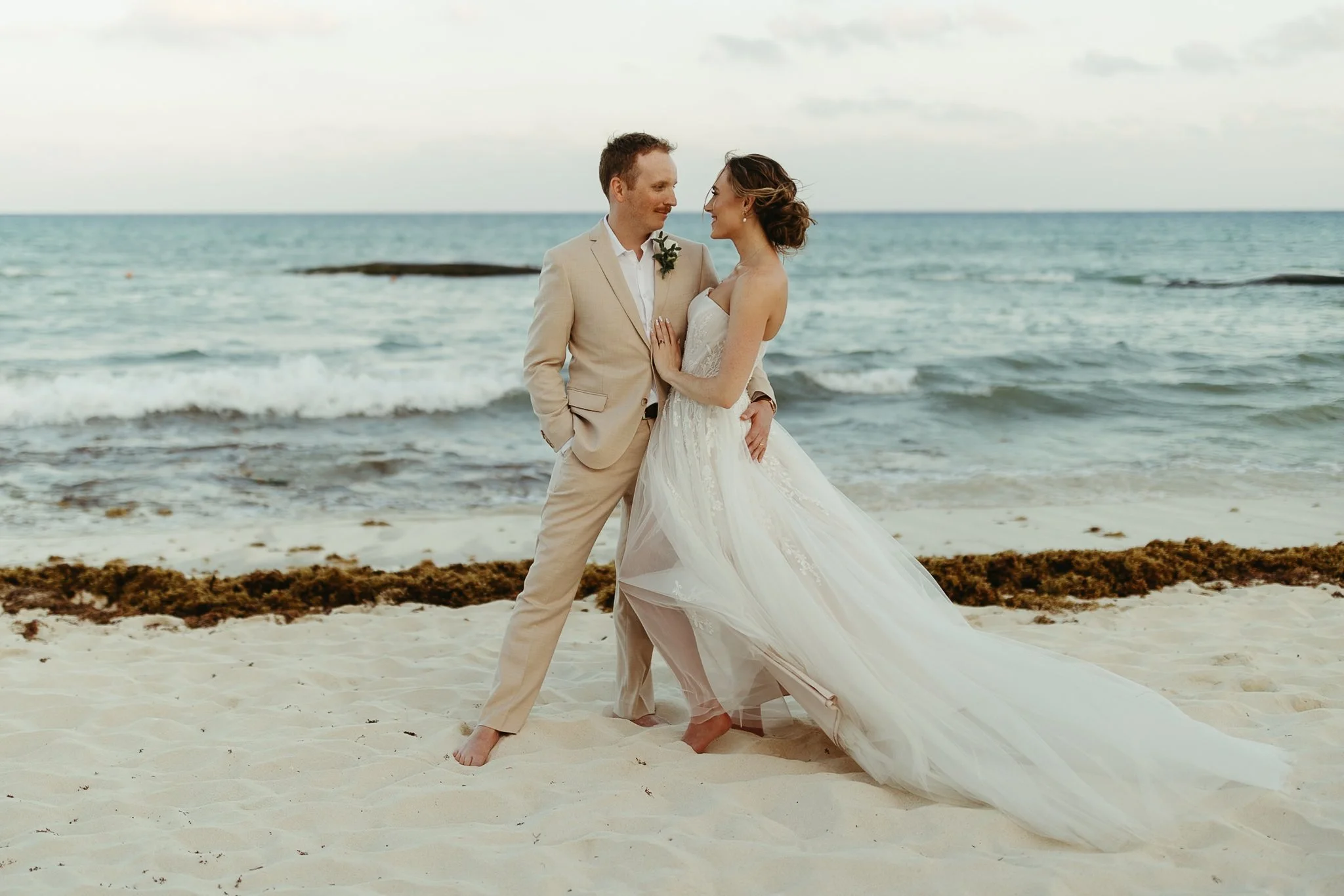 A newlywed couple in wedding attire standing barefoot on the beach near the ocean, at The Fives Beach Resort in Playa Del Carmen, Mexico gazing at each other.