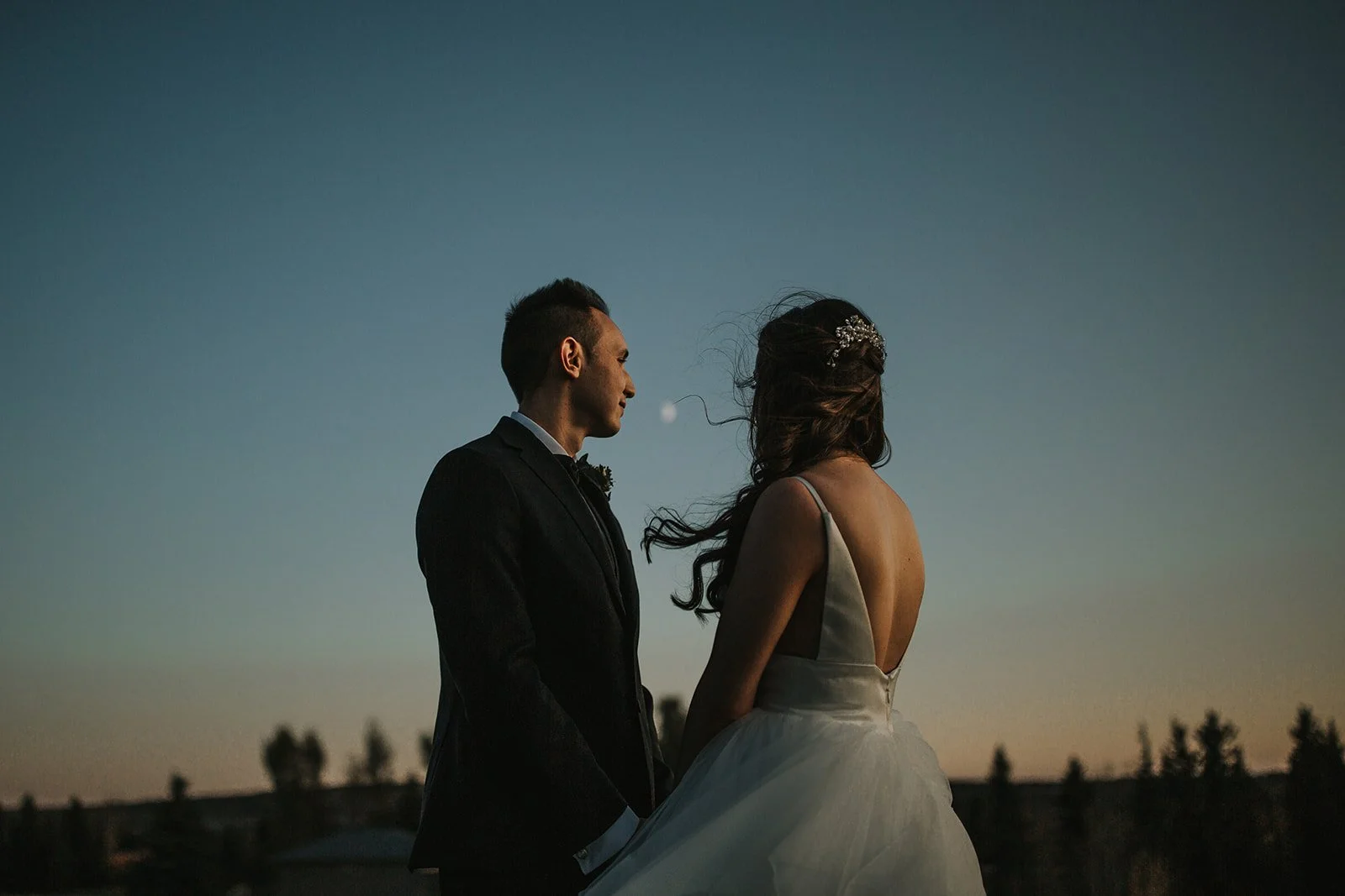 Bride and groom stand outside at sunset with the full moon in the sky between them