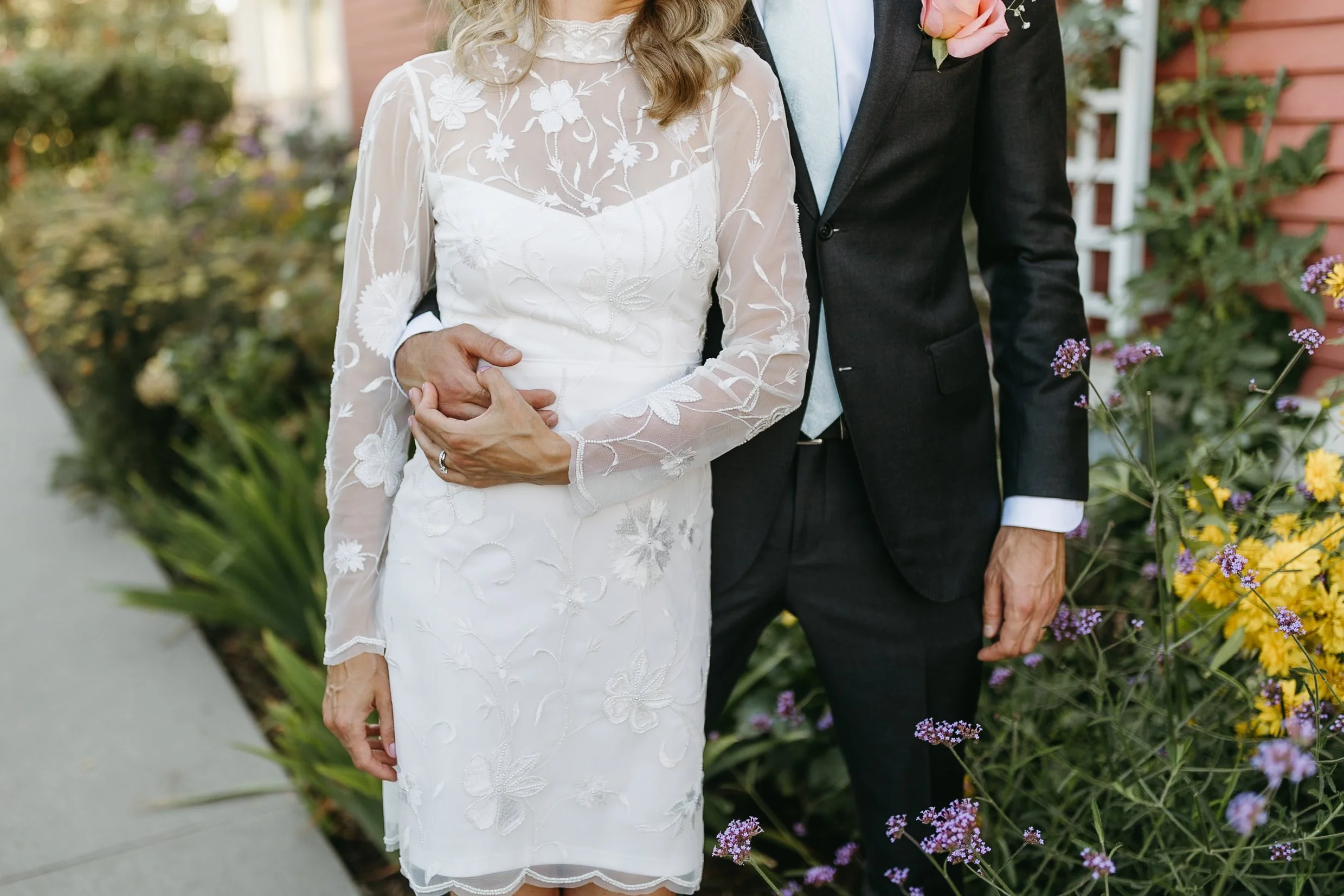 A bride and groom standing together outside during their wedding at Deane House in Calgary, the bride in a white lace dress and the groom in a black suit with a light blue tie, surrounded by colourful flowers and greenery.